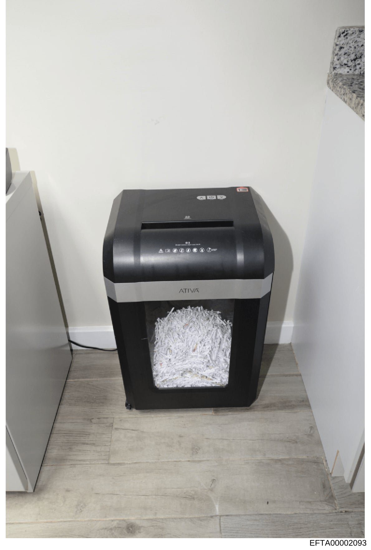 Photograph of a black and silver Ativa brand paper shredder containing shredded documents. The shredder is positioned on a light wood-tone floor against white walls. This image is documented evidence, likely taken during a search or seizure related t