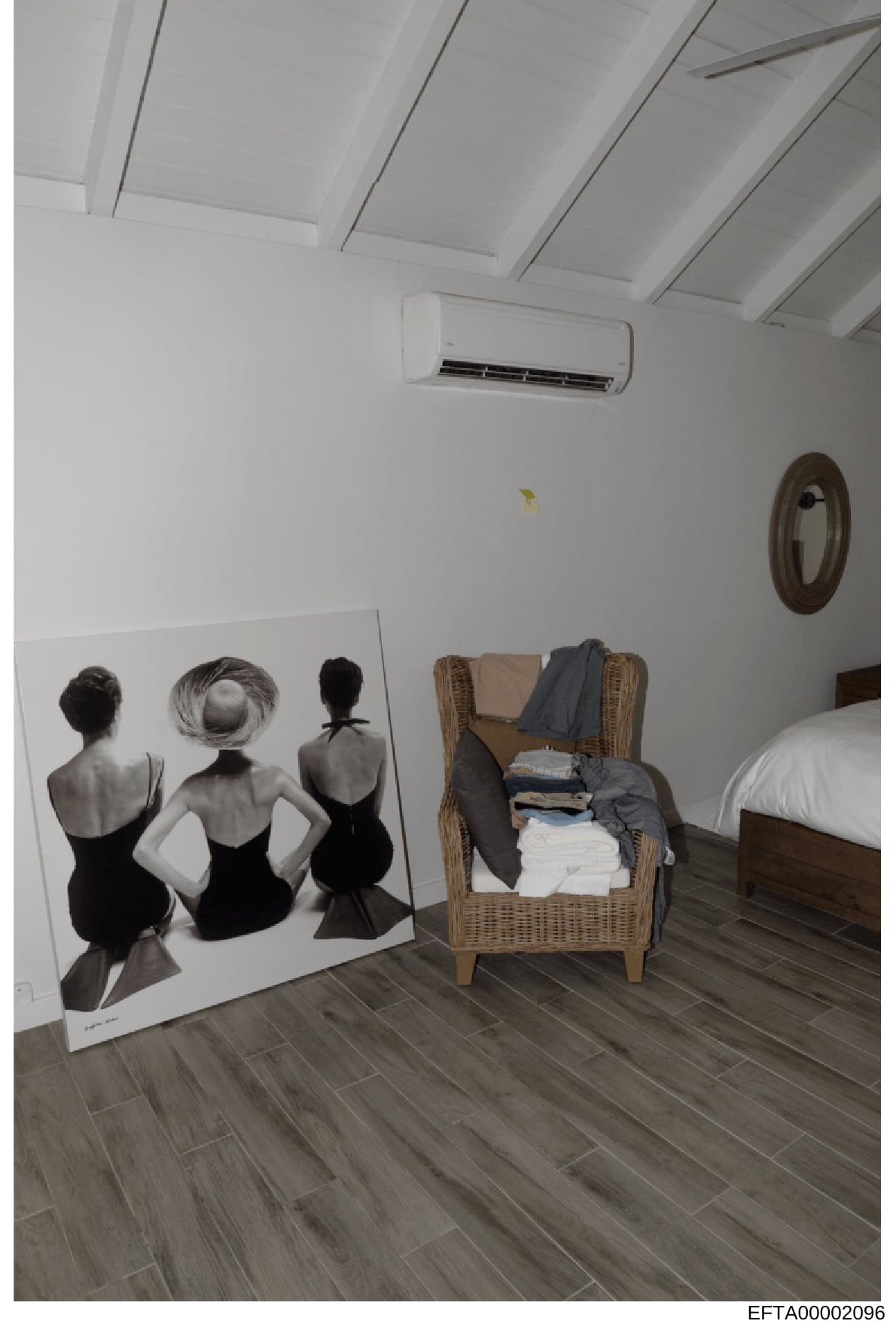 This is a photograph of a bedroom interior featuring a large artistic print depicting three female figures from behind, a wicker chair with folded clothing and textiles, a bed with white bedding, and modern amenities including an air conditioning uni