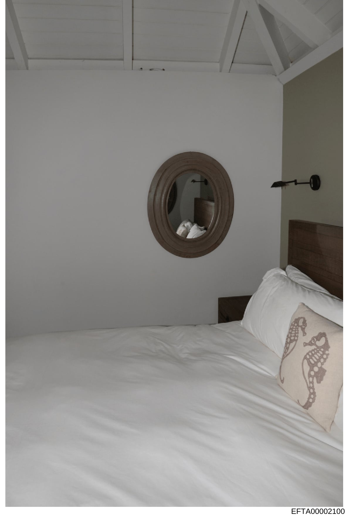 This is a photograph of a bedroom interior showing a made bed with white linens and decorative pillows, a round framed mirror on the wall, and a modern wall sconce. The image appears to be documentation of property interior details, possibly from one