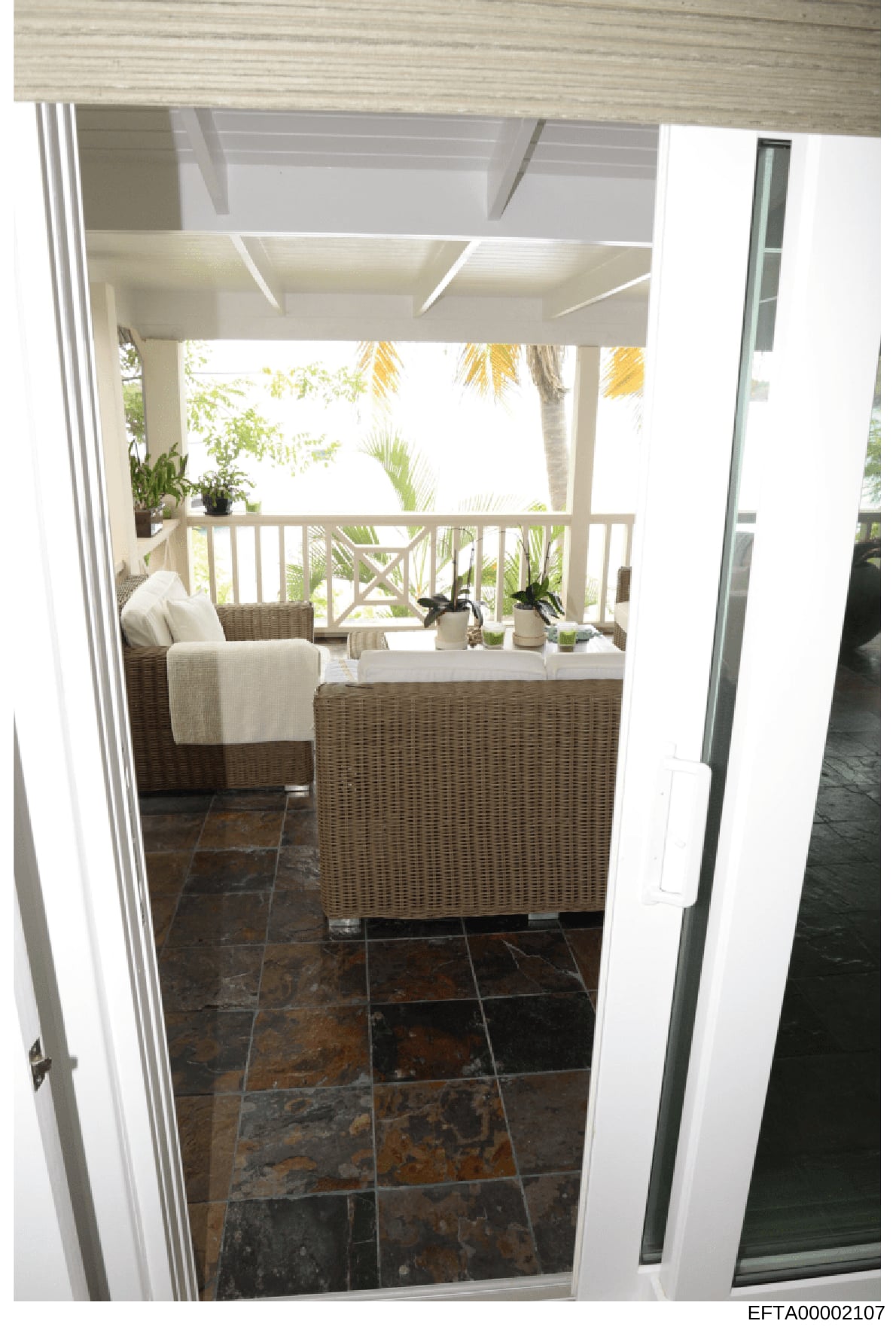 This is a photograph of a luxurious covered outdoor living space, showing a screened or covered porch with high-end wicker furniture, potted plants, and views to tropical landscaping. The image appears to be from an interior design or property docume