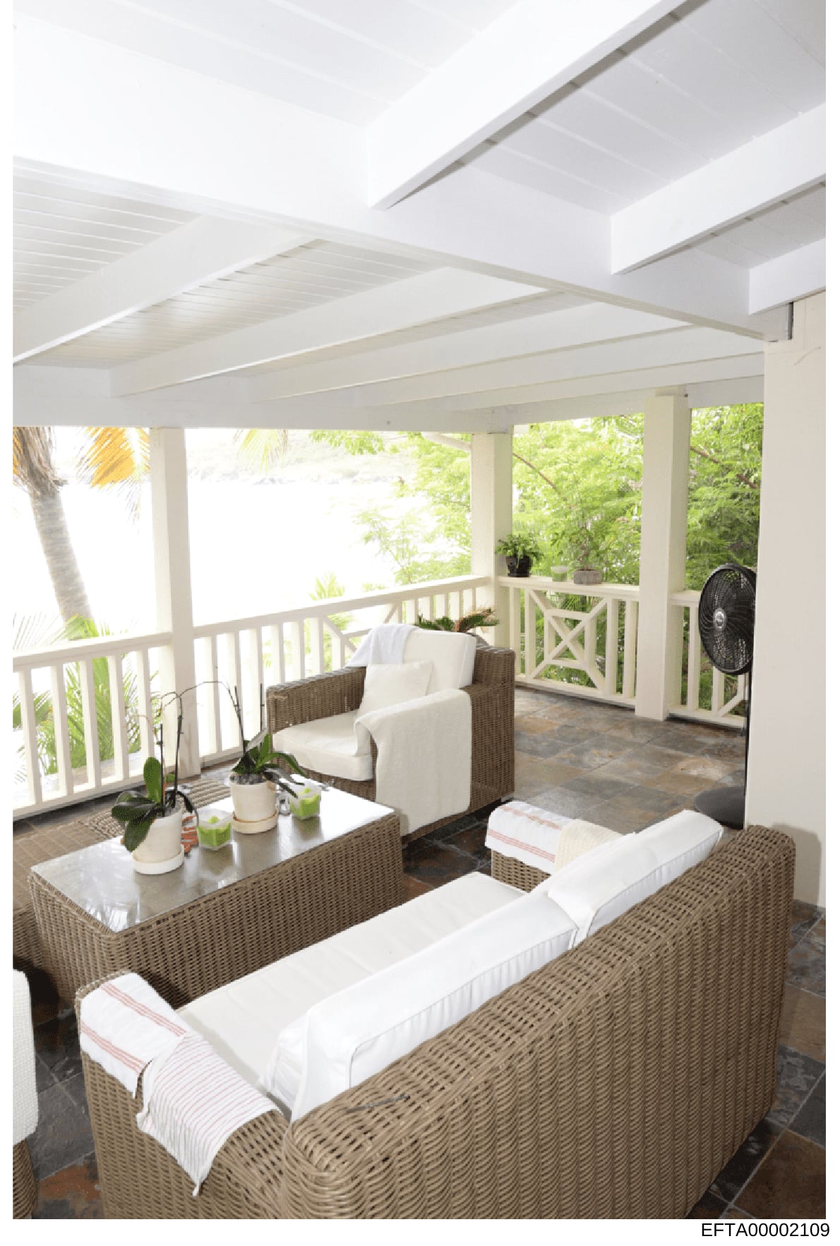 This is a photograph of an elegant covered outdoor space featuring wicker furniture with white cushions, potted plants, and views of tropical landscaping. The image shows a well-maintained residential porch or veranda with decorative white railings,