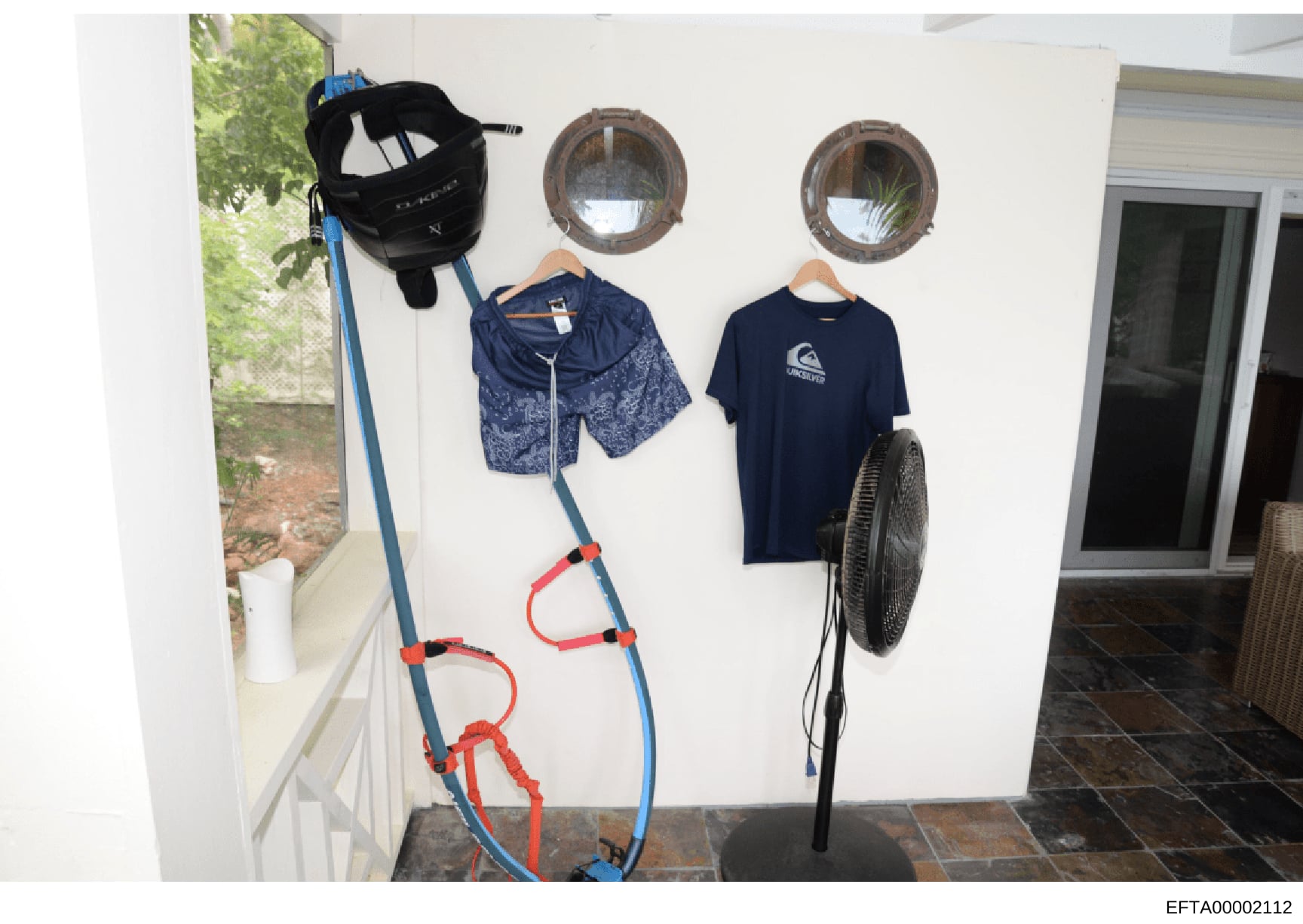 This photograph shows an interior room, likely a mudroom or entryway, displaying various sports and recreational equipment hung on white walls. The image includes water sports gear (wakeboard, life vests), athletic clothing, and household items. This