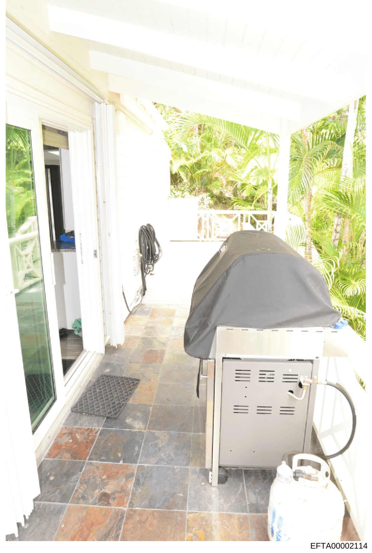 This is an evidence photograph showing an interior covered porch or screened area of a property. The image depicts a tiled floor, white framing, tropical vegetation visible through openings, a covered grill or similar outdoor equipment, coiled hoses,