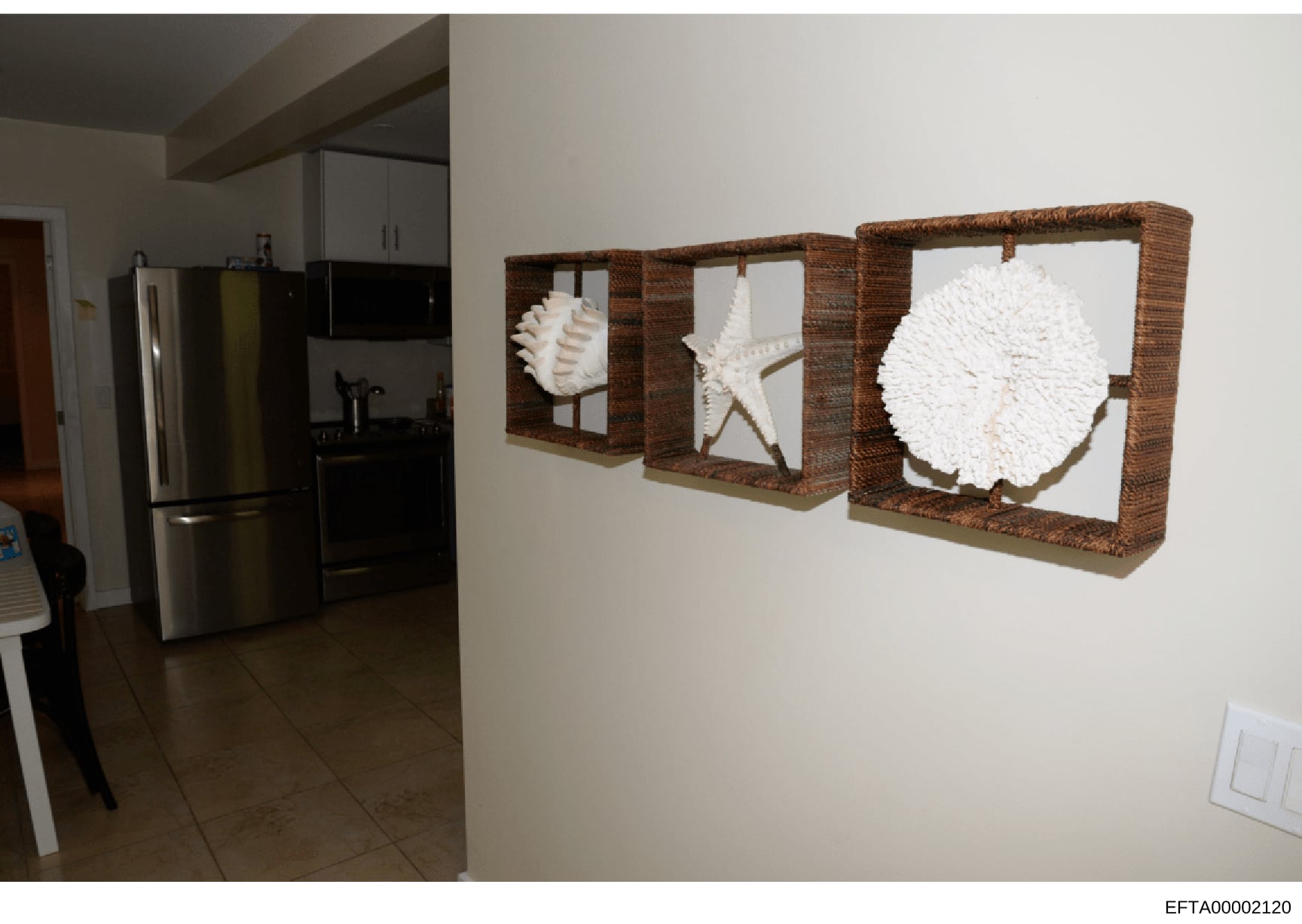 This is a photograph of a residential kitchen and adjacent room showing modern appliances, cabinetry, and three framed nautical-themed wall art pieces featuring white shells and starfish in woven frames. The image appears to be evidence documentation
