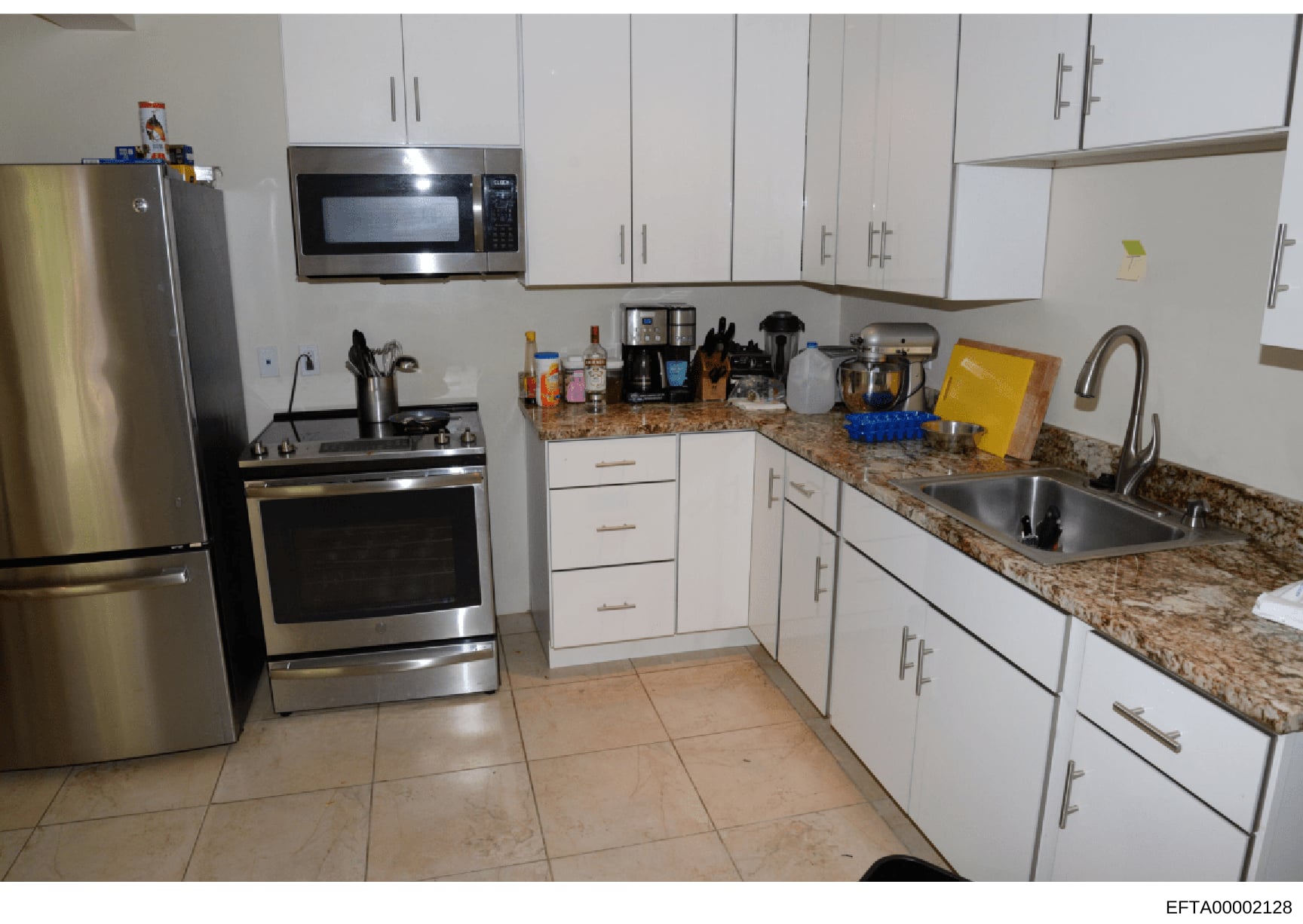 This is a photograph of a modern residential kitchen featuring white cabinetry, granite countertops, and stainless steel appliances. The image appears to be evidence documentation from a property investigation, showing the kitchen's layout, furnishin