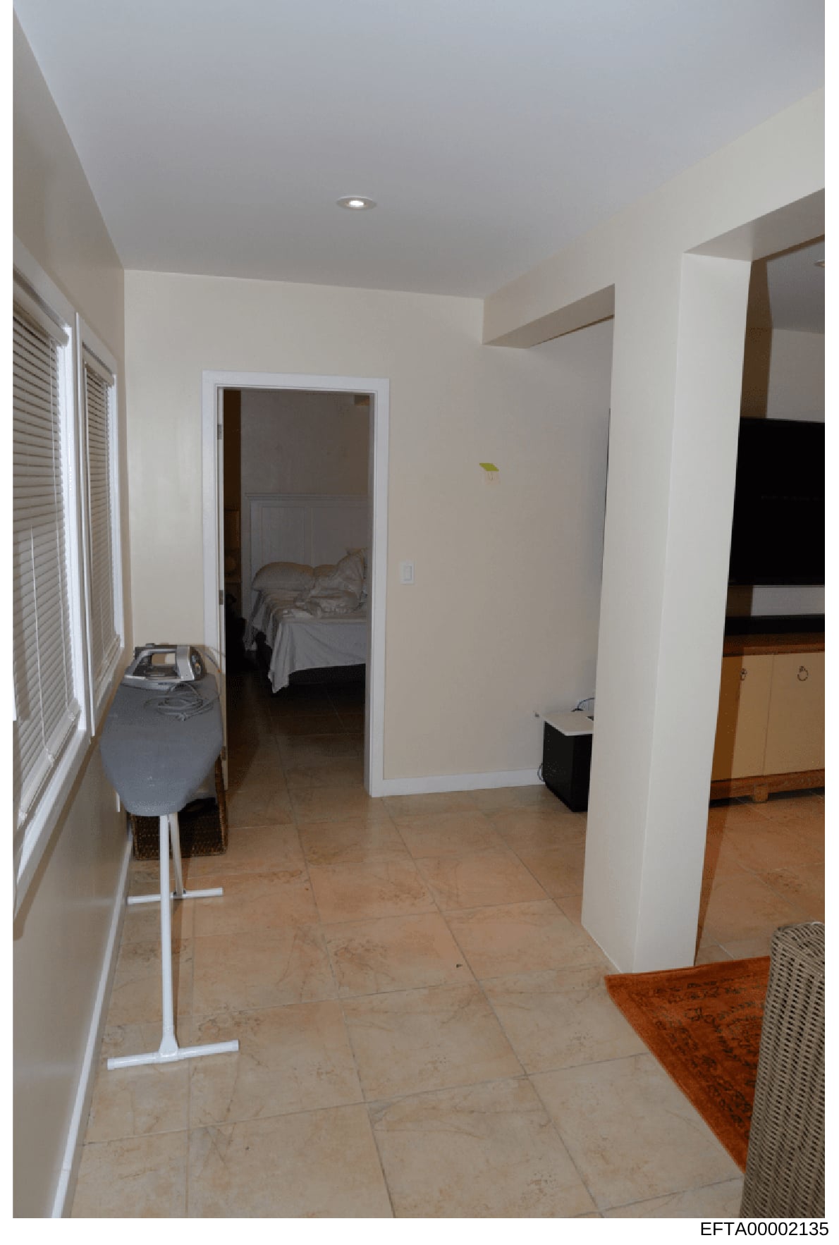 This is a photograph of an interior hallway or entryway of a residential property, showing tile flooring, white walls, an open doorway to a bedroom, and various household items. The image appears to be part of property documentation, possibly related