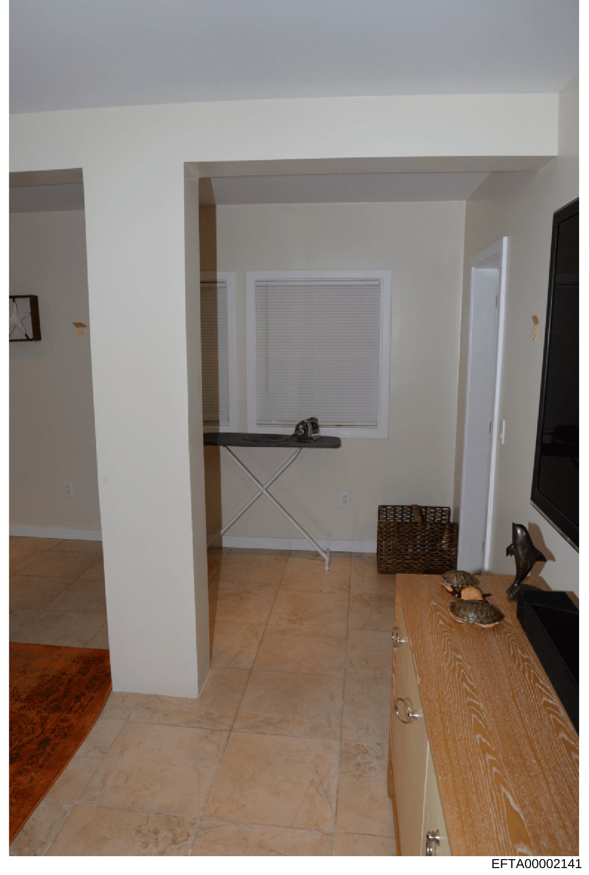 This is an evidence photograph showing an interior room with white walls, beige/tan tile flooring, a window with white blinds, a black shelving unit, and various household items. The image appears to be documentation of a residential space, possibly 