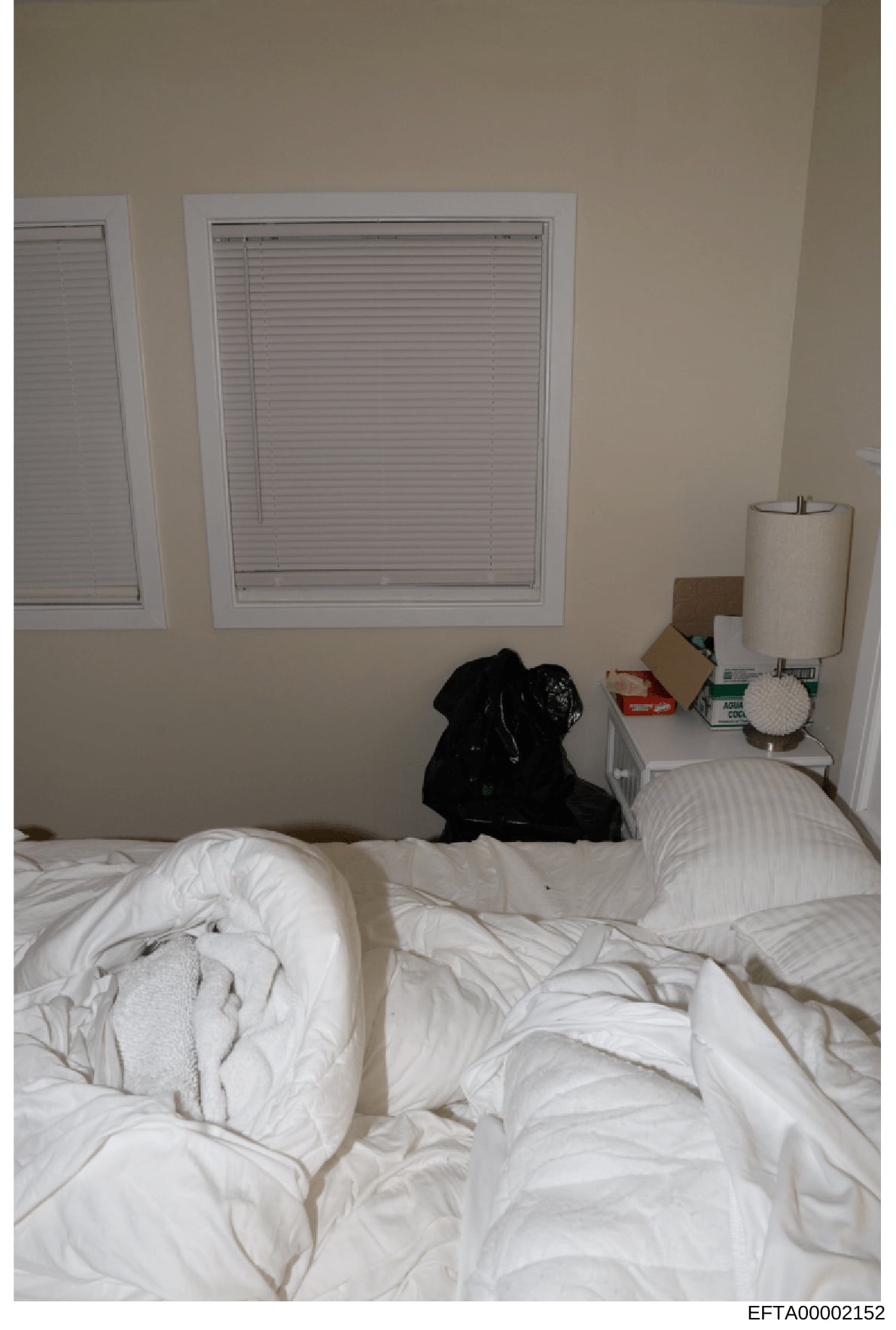 This is a photograph of a residential bedroom showing an unmade bed with white linens, two windows with white blinds, and personal items on a nightstand including a paper towel holder and miscellaneous boxes. The image appears to be evidence document