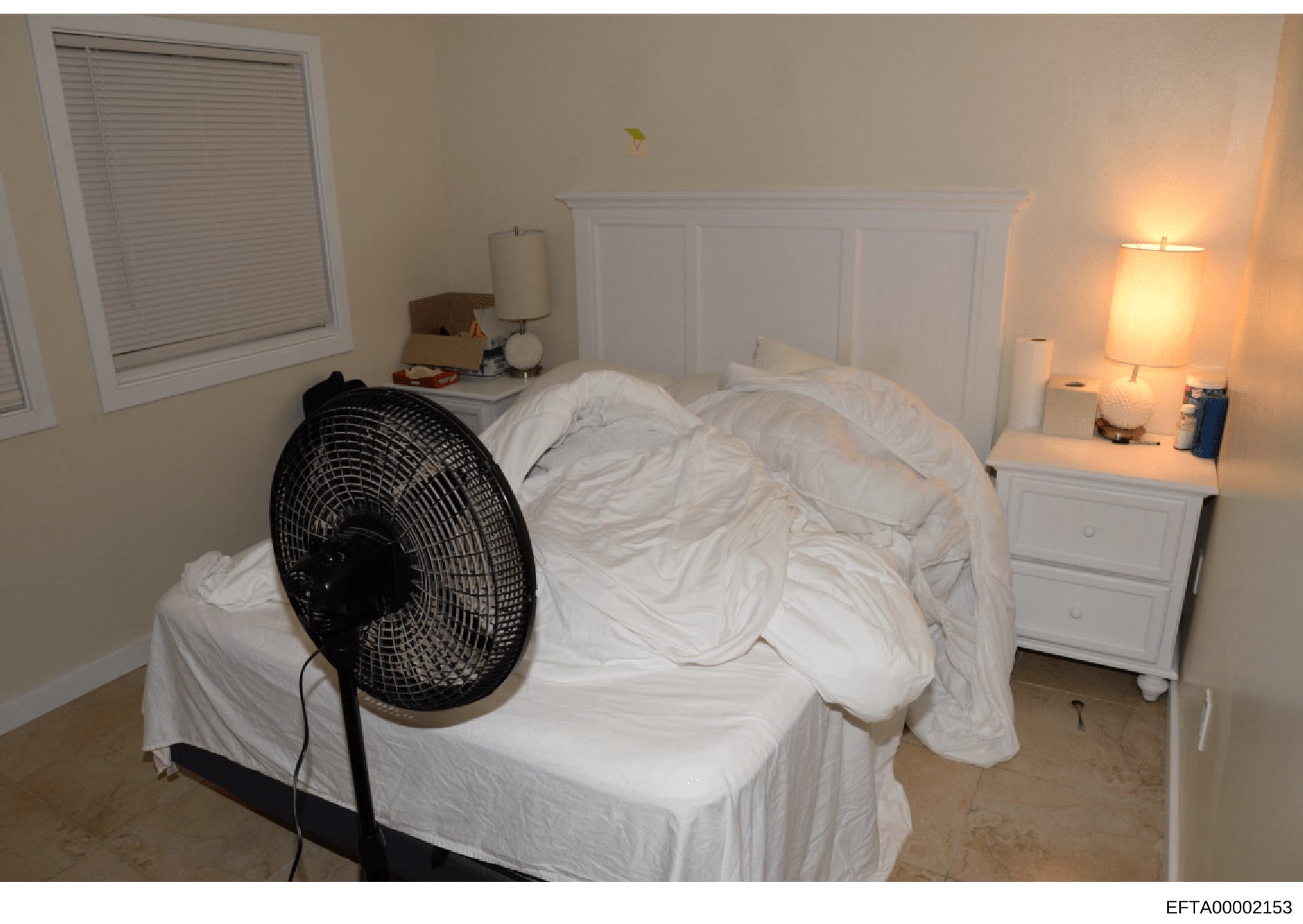 This is an evidence photograph of a bedroom interior, showing a bed with white linens, a black pedestal fan, white paneled headboard, nightstands with lamps, a window with blinds, and basic furnishings. The image appears to be documentation of a resi