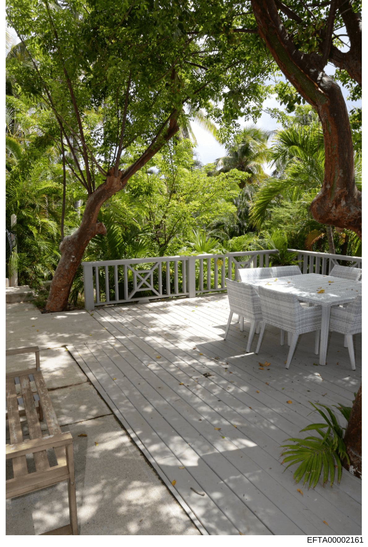 This photograph shows an exterior deck or patio area featuring white wicker furniture, a dining table, and extensive tropical landscaping with mature trees and palm vegetation. The image appears to be documentation of a residential property, likely p