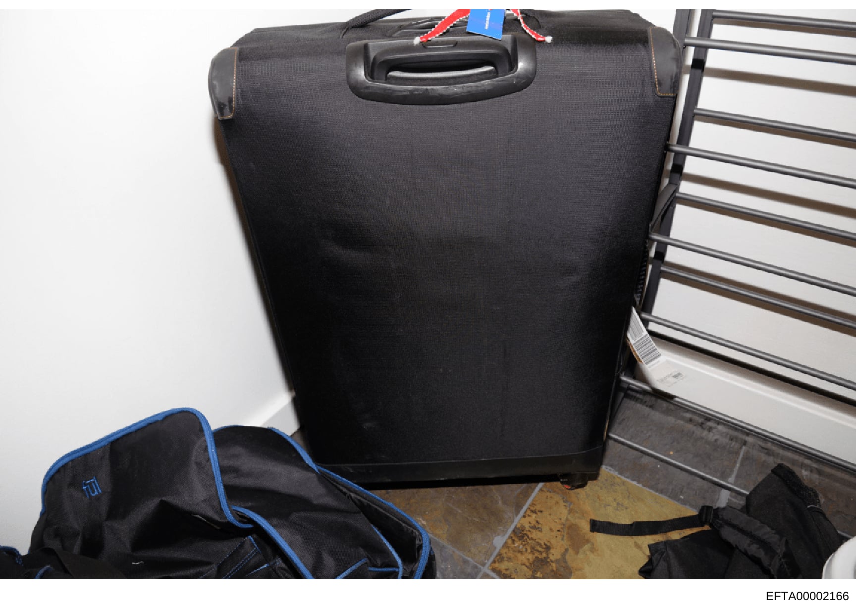 This is an evidence photograph showing a black rolling suitcase with a blue ID tag and red/white cord attached to the handle. The suitcase appears to be stored or photographed in an indoor setting against a white wall, with metal railings visible. Th