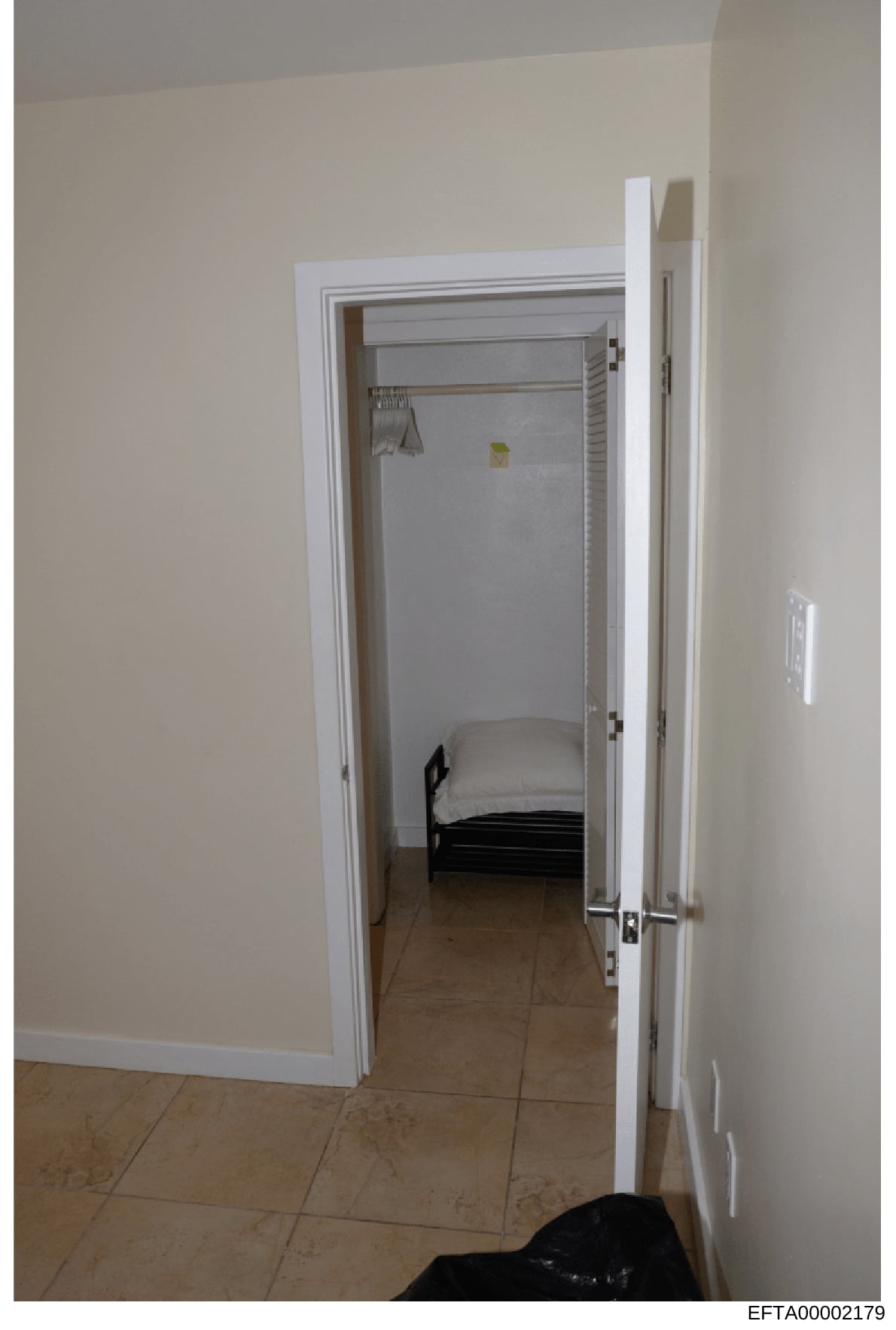 This is an evidence photograph showing the interior of a small bedroom, viewed through an open doorway. The room contains a single bed with black frame and white bedding, white walls, tile flooring, and basic furnishings. The photograph appears to be
