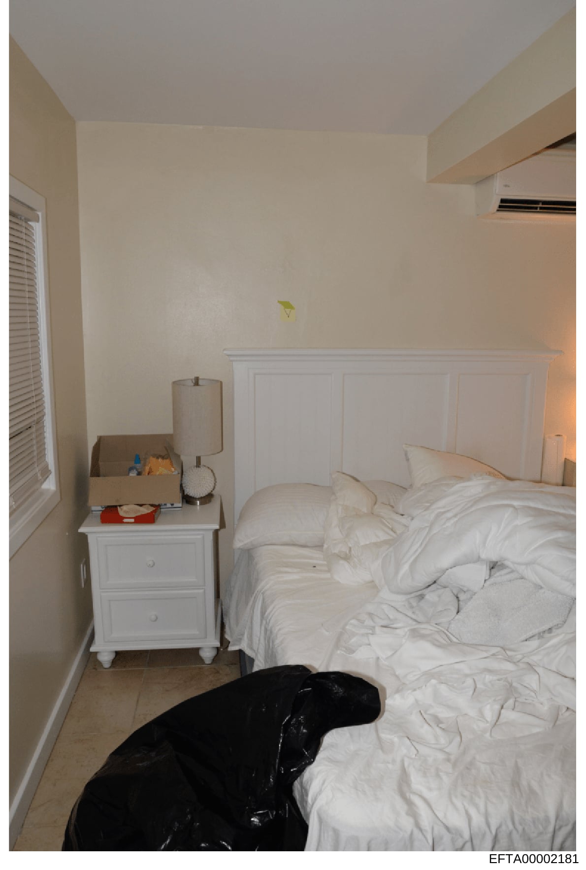 This is a photograph of a simple bedroom interior showing a bed with white linens, white nightstand, white headboard paneling, and basic furnishings. The image appears to be evidence documentation from the investigation, with a Bates number indicatin