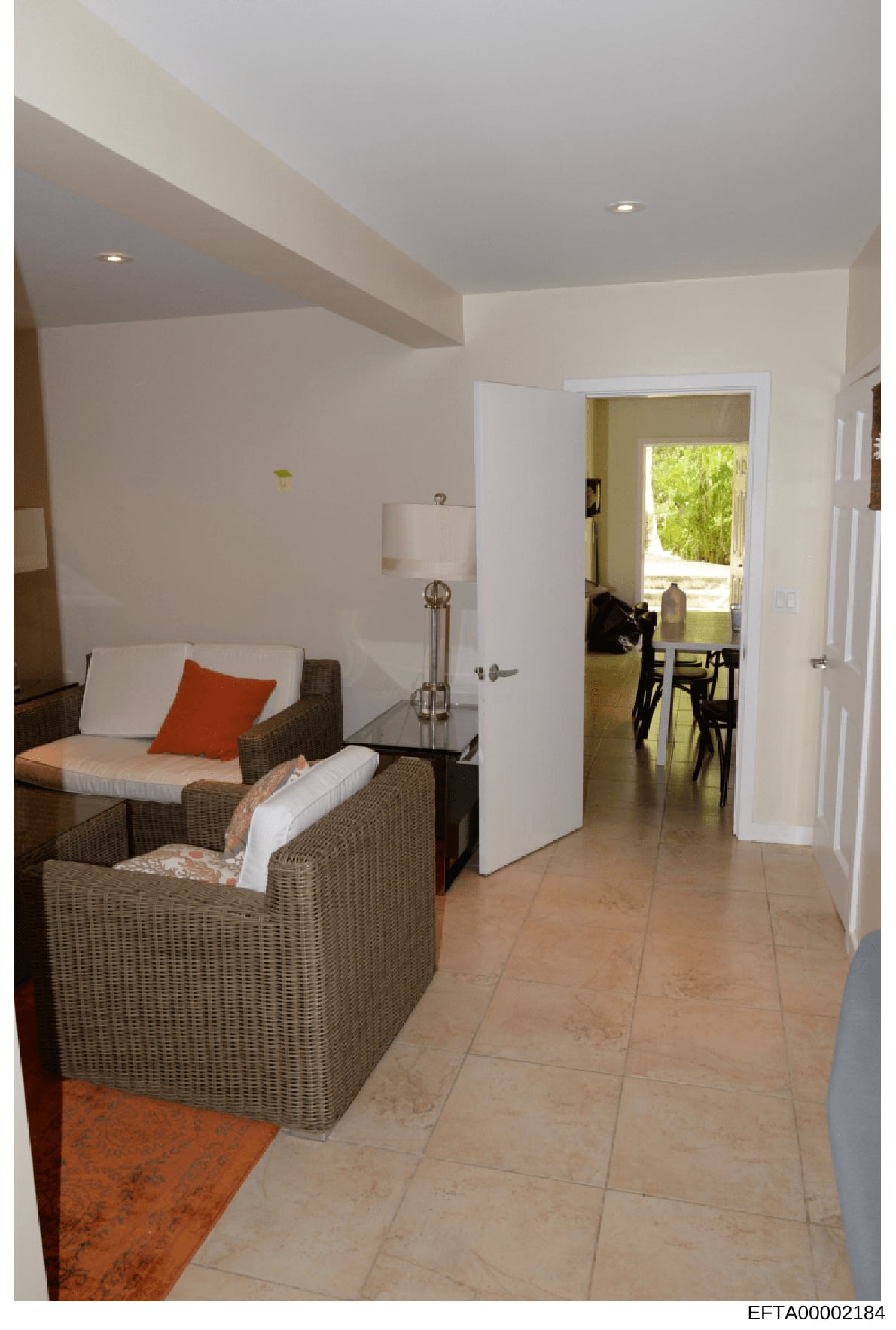 This is a photograph of a modern interior room, likely a bedroom or suite, showing contemporary furnishings and design. The image depicts woven rattan chairs with white cushions and an orange accent pillow, a side table with lamp, white walls with ex