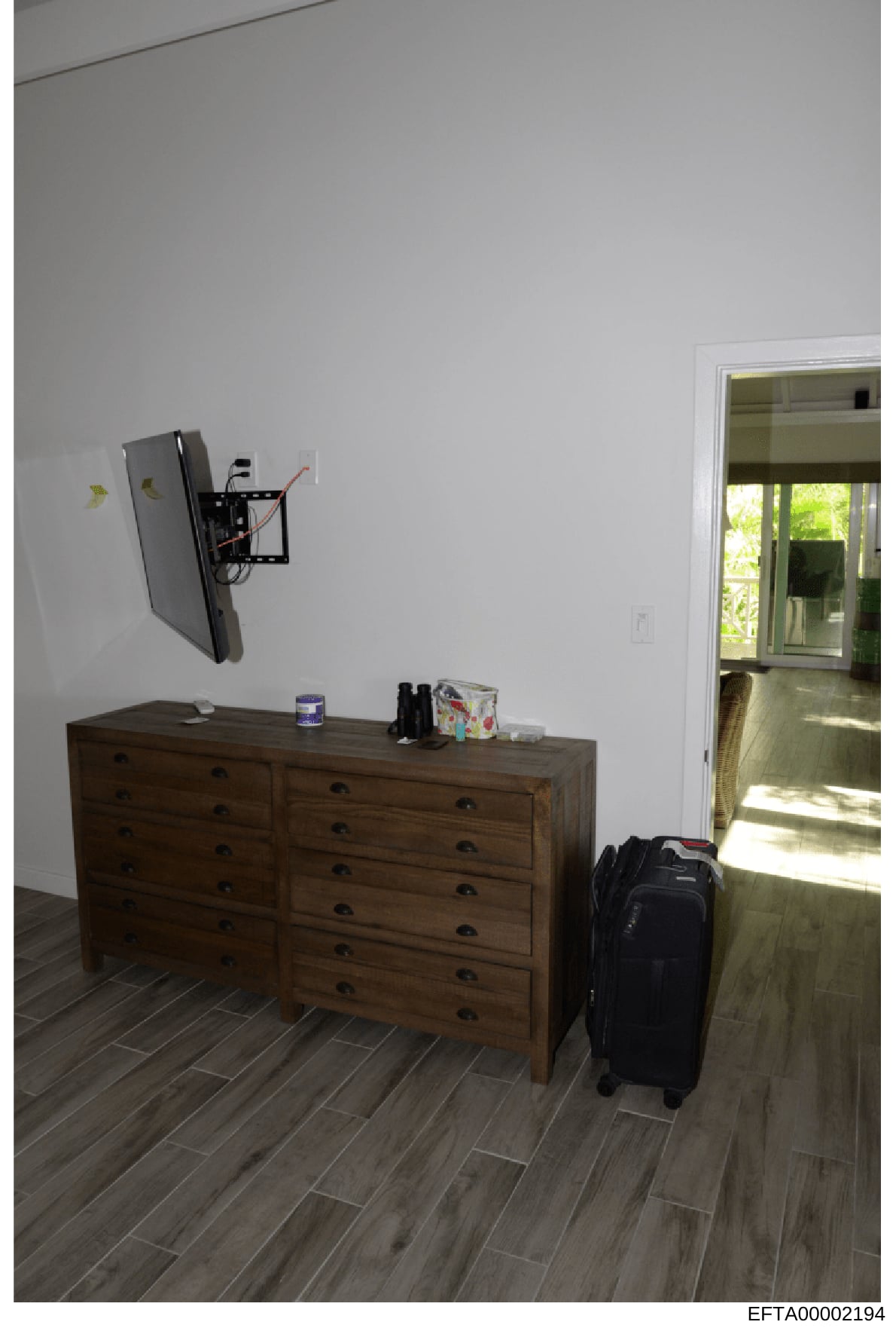 This is a photograph of a residential bedroom interior showing a wooden dresser, wall-mounted flat-screen television with bracket, and glass door opening to an outdoor area. The image appears to be documentary evidence from a property inspection or i