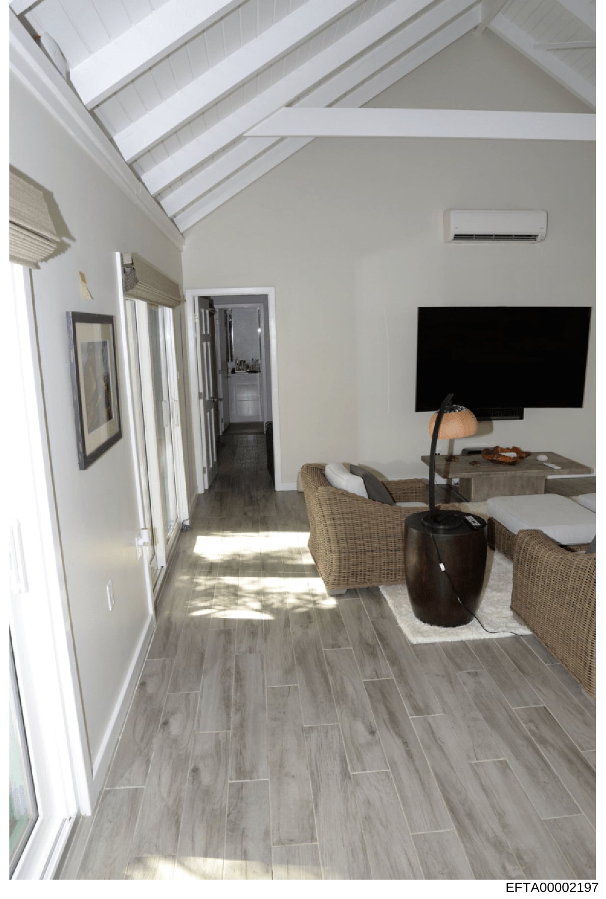 This photograph shows the interior of a modern residential room, featuring contemporary design elements including vaulted ceilings with skylights, light wood-look flooring, wicker furnishings, and a wall-mounted flat-screen television. The image appe