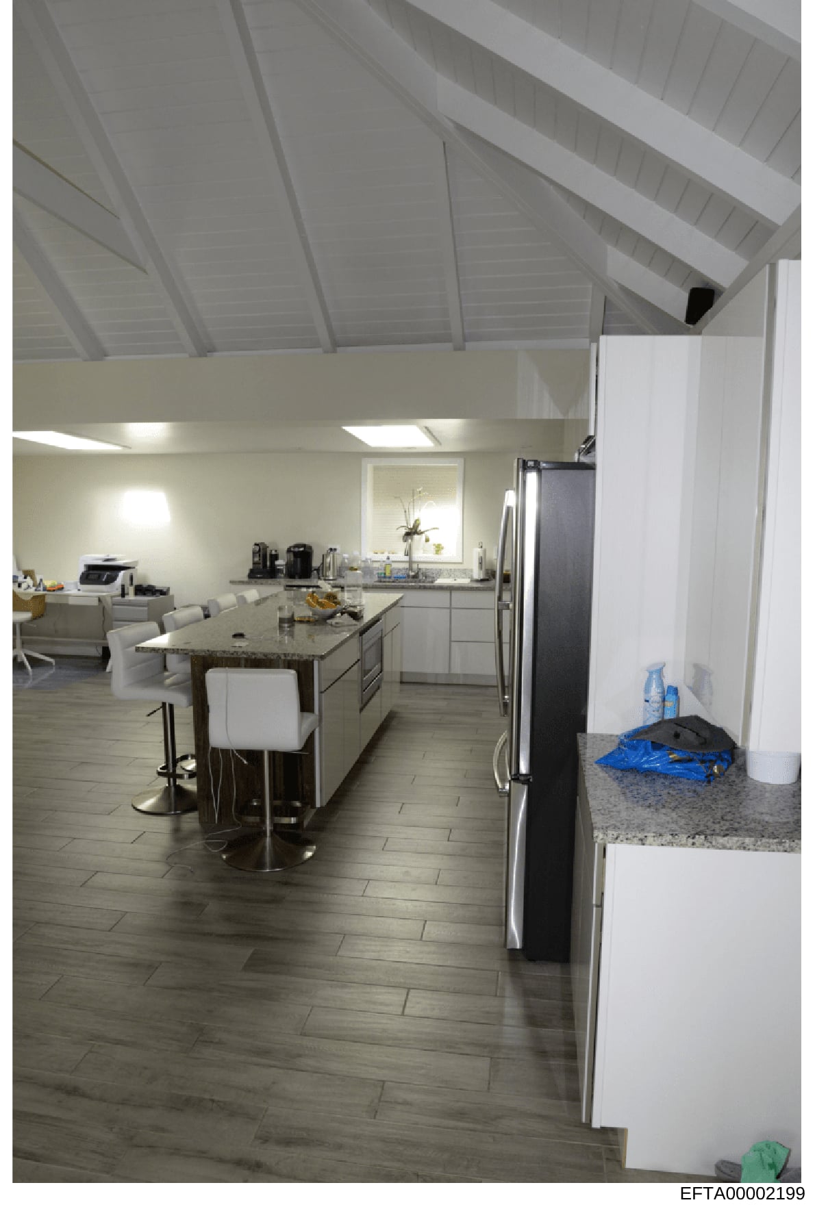 This photograph shows a modern kitchen and connected living area, featuring contemporary design elements including white cabinetry, stainless steel appliances, an island with seating, and vaulted ceilings. The image appears to be documentation of a r