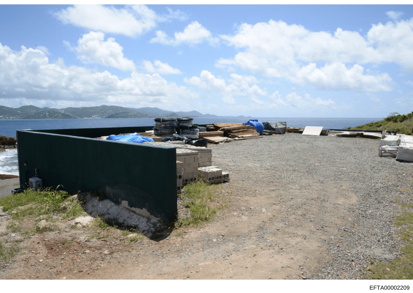 Photograph of a waterfront storage or work area showing various maritime equipment, construction materials, and storage structures along a rocky coastline. The image depicts a property with ocean views and mountains in the background, containing orga