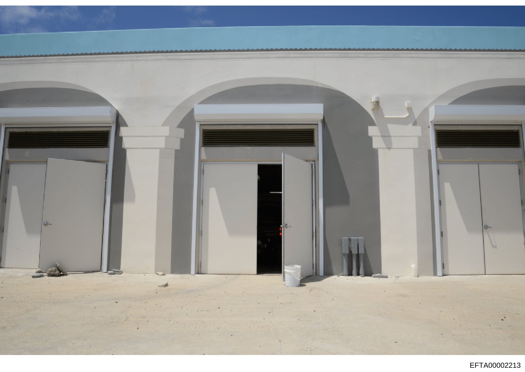 This photograph shows the exterior of a white concrete building structure located on a beach, featuring multiple arched entrances with white doors. The building has a simple, modern design with a turquoise roof visible at the top. The image appears t