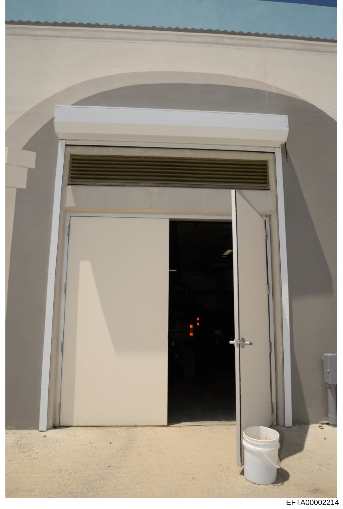 This photograph shows an arched interior doorway with white trim and a closed white door. The door is partially open, revealing a darkened interior space with red lights visible inside. A white bucket is positioned on the sandy floor in front of the 