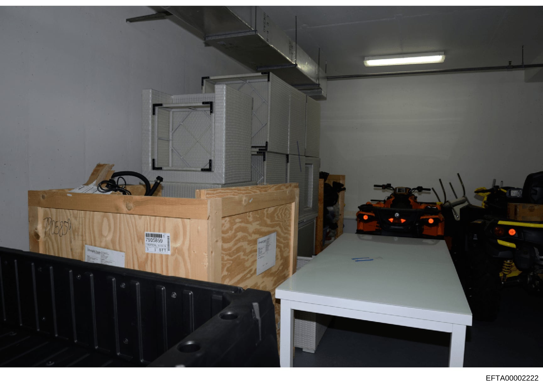 This photograph shows the interior of a storage or warehouse facility containing multiple wooden shipping crates with barcode labels, various equipment including orange and black machinery or devices, metal shelving, industrial lighting, and what app