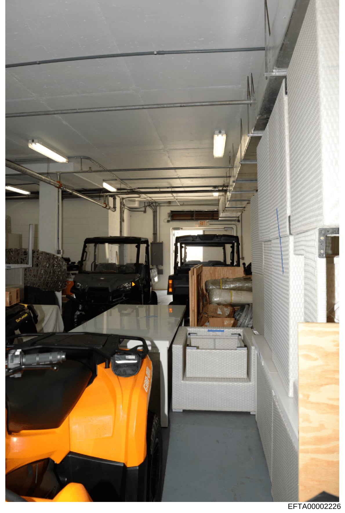 This photograph shows the interior of a large warehouse or storage facility containing multiple vehicles including an orange forklift in the foreground and at least two black all-terrain vehicles or utility vehicles in the background. The space appea