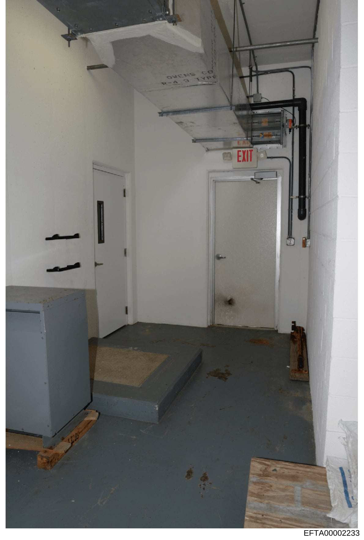 This is an evidence photograph showing the interior of a basement or utility room space, featuring white walls, exposed electrical and HVAC systems, an illuminated red EXIT sign, and various equipment and materials on the floor. The image appears to