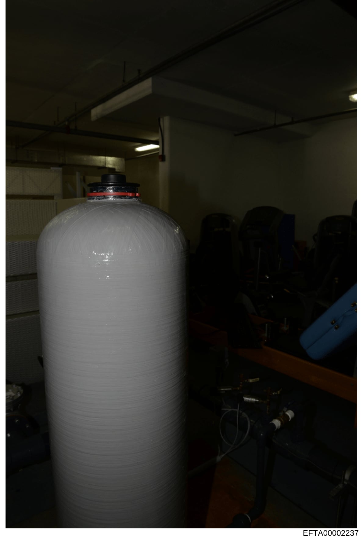 Photograph of a large white cylindrical storage tank or water filtration system in an indoor facility. The equipment appears to be industrial or commercial in nature, featuring a red valve assembly at the top and black piping connections. The image w