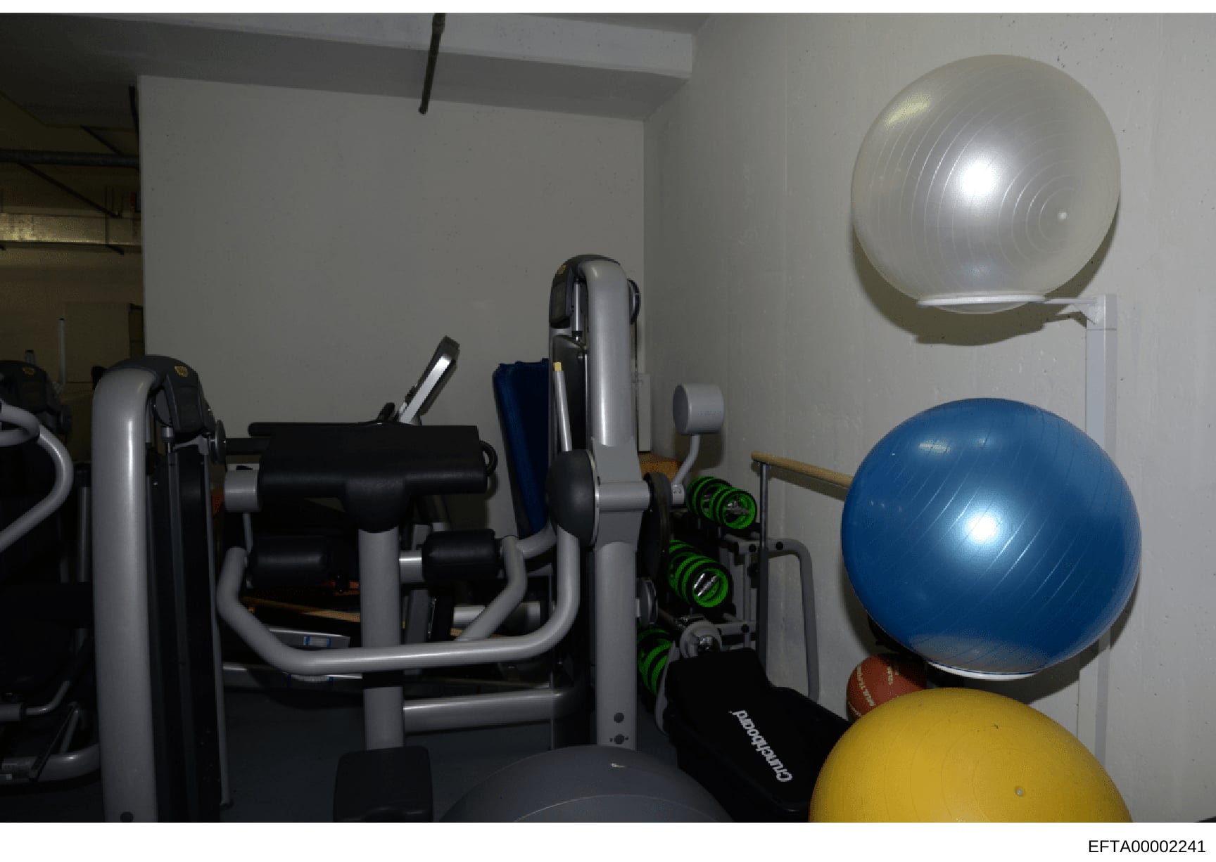 This is a photograph of a residential gym or fitness room showing various exercise equipment and accessories. The image captures exercise machines, stability balls, dumbbells, and other workout gear stored in what appears to be a basement or dedicate