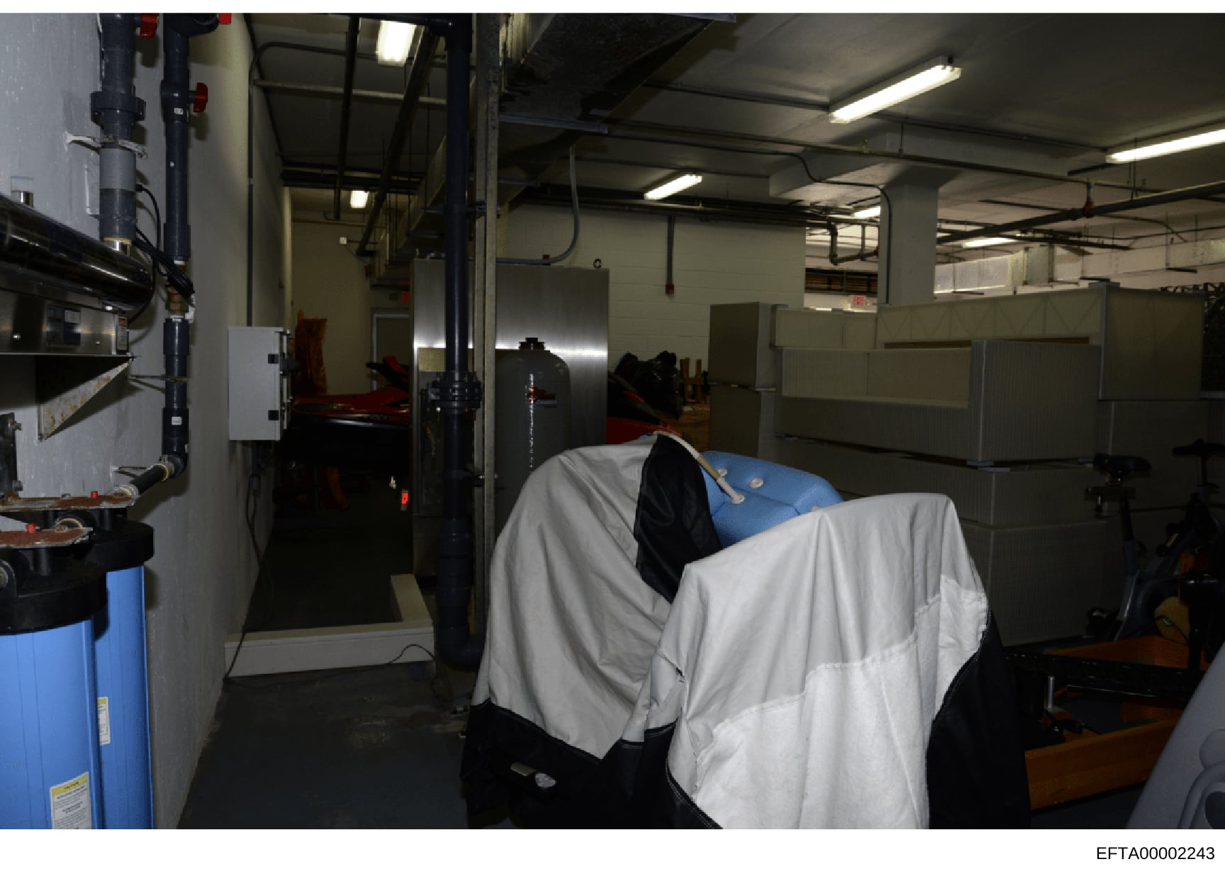 This photograph shows an industrial or basement-level space with utility equipment, mechanical systems, and storage areas. The image captures what appears to be a service or mechanical room with fluorescent lighting, exposed piping, metal storage con