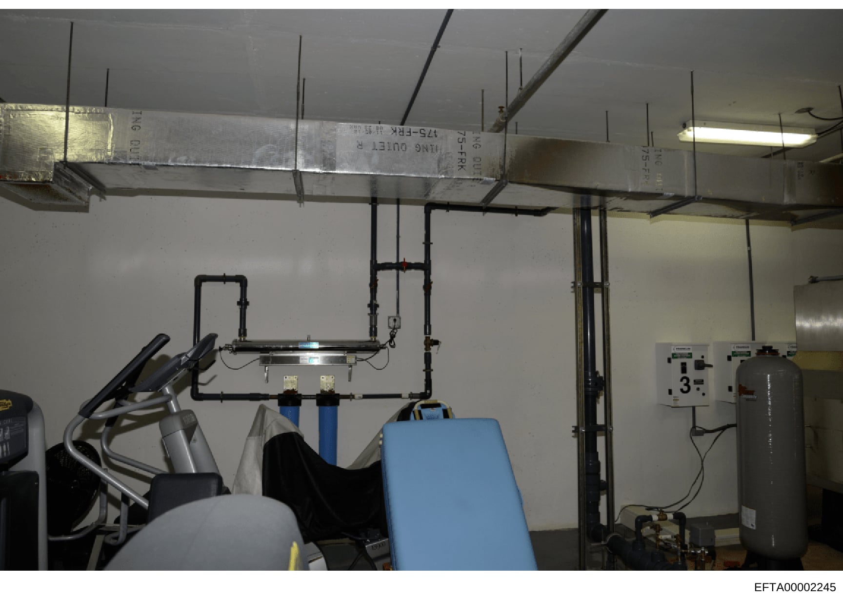 This photograph shows an interior room with exposed mechanical systems including ductwork, piping, and HVAC equipment mounted on walls and ceiling. A blue piece of exercise or medical equipment is visible in the foreground. The image appears to be do