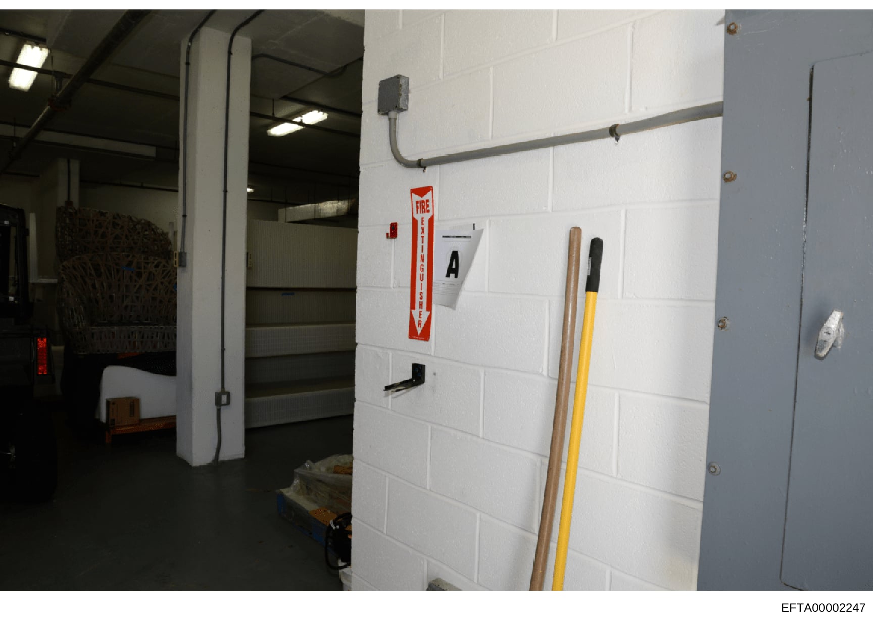 This photograph shows an interior utility or storage space with white cinder block walls, basic infrastructure, and safety equipment. The room contains a fire extinguisher sign, electrical conduit, and various tools. The image appears to be evidence 