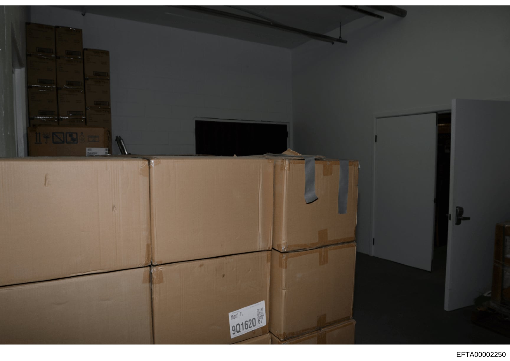 This photograph shows an interior storage or warehouse space containing multiple sealed cardboard boxes stacked against a wall, with shelving units visible in the background and a television monitor on top of boxes. The image appears to be evidence d