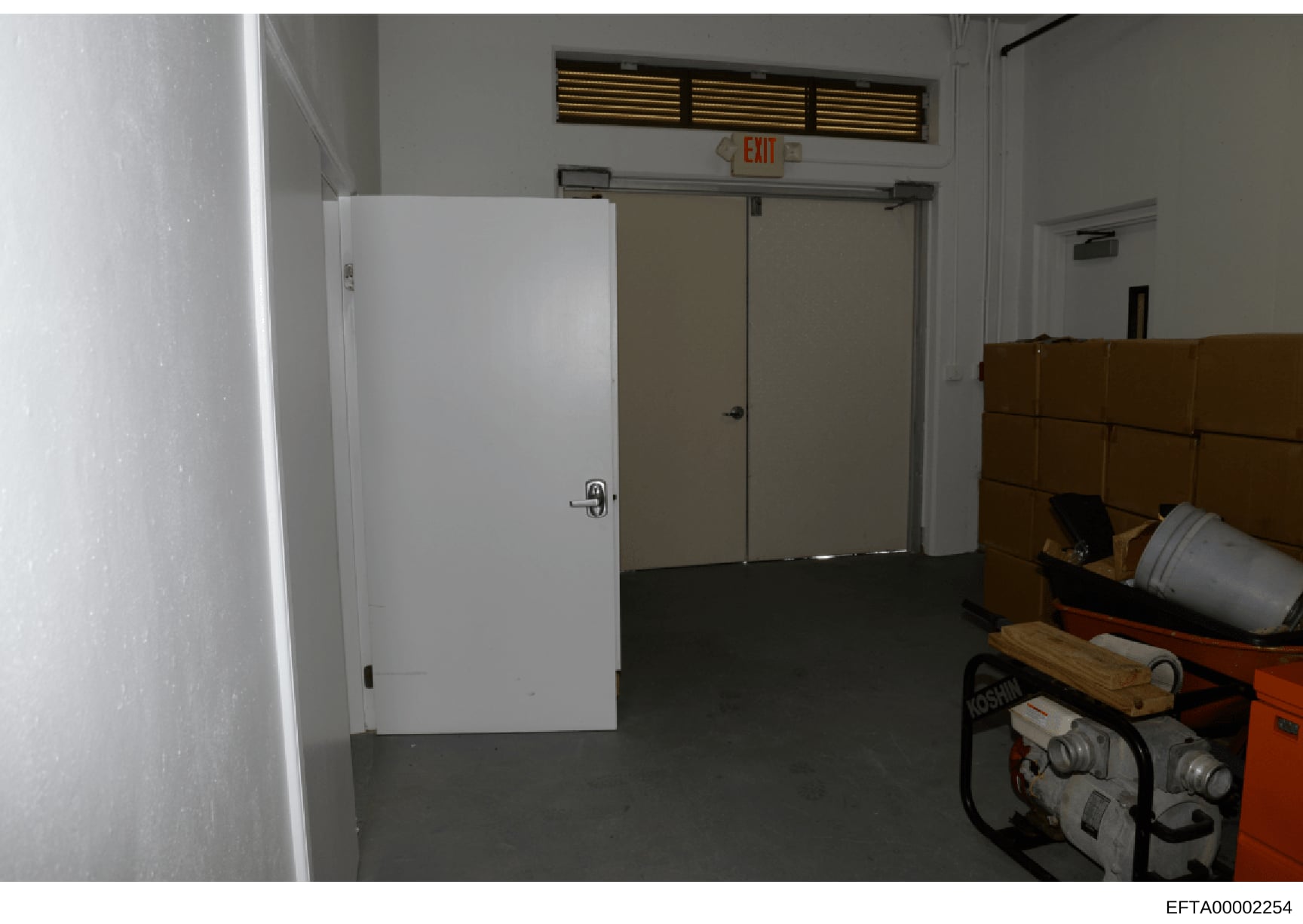 This is a photograph of an interior room or basement space, showing white doors, fluorescent lighting, an EXIT sign, and various equipment and storage items including boxes and tools. The image appears to be evidence documentation from a property sea