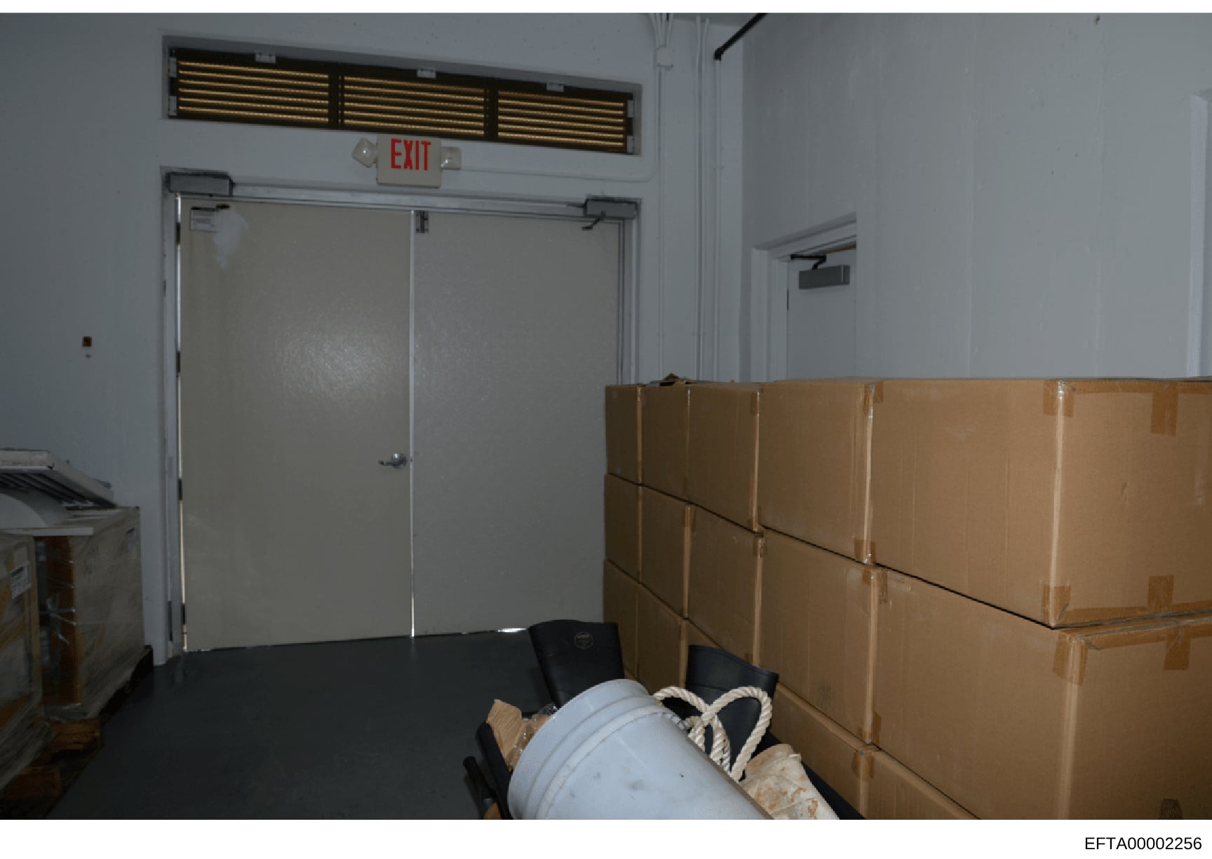 This is an evidence photograph showing an interior storage or warehouse space with stacked cardboard boxes, a white pipe or container, and an EXIT sign visible. The image documents the physical condition and contents of what appears to be a storage a