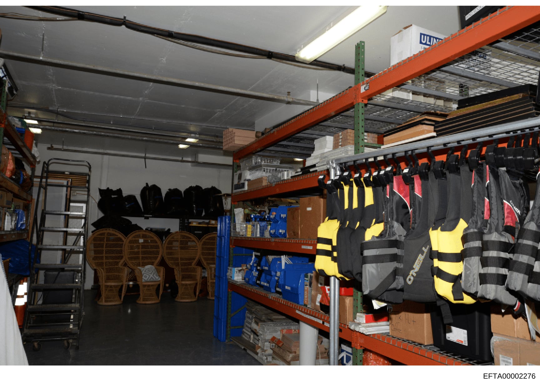 This is a photograph of an industrial warehouse or storage facility containing various equipment and supplies. The image shows organized storage with orange industrial shelving units holding life jackets, wetsuits, helmets, paddles, and other water s