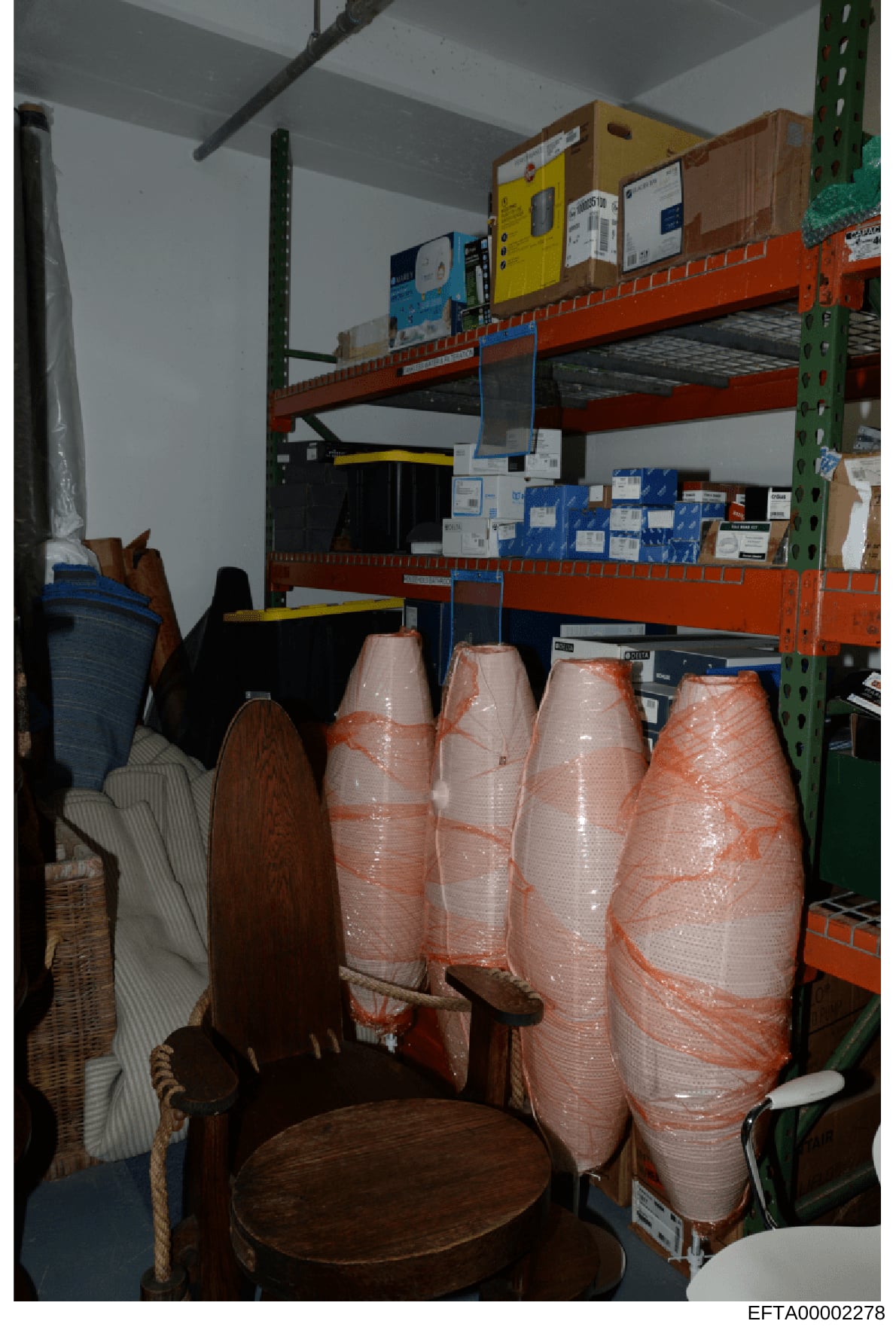 This photograph shows an evidence storage room or warehouse space containing wrapped furniture items, cardboard boxes, and various stored goods on industrial shelving. The image appears to be documentation of seized or stored property, with multiple