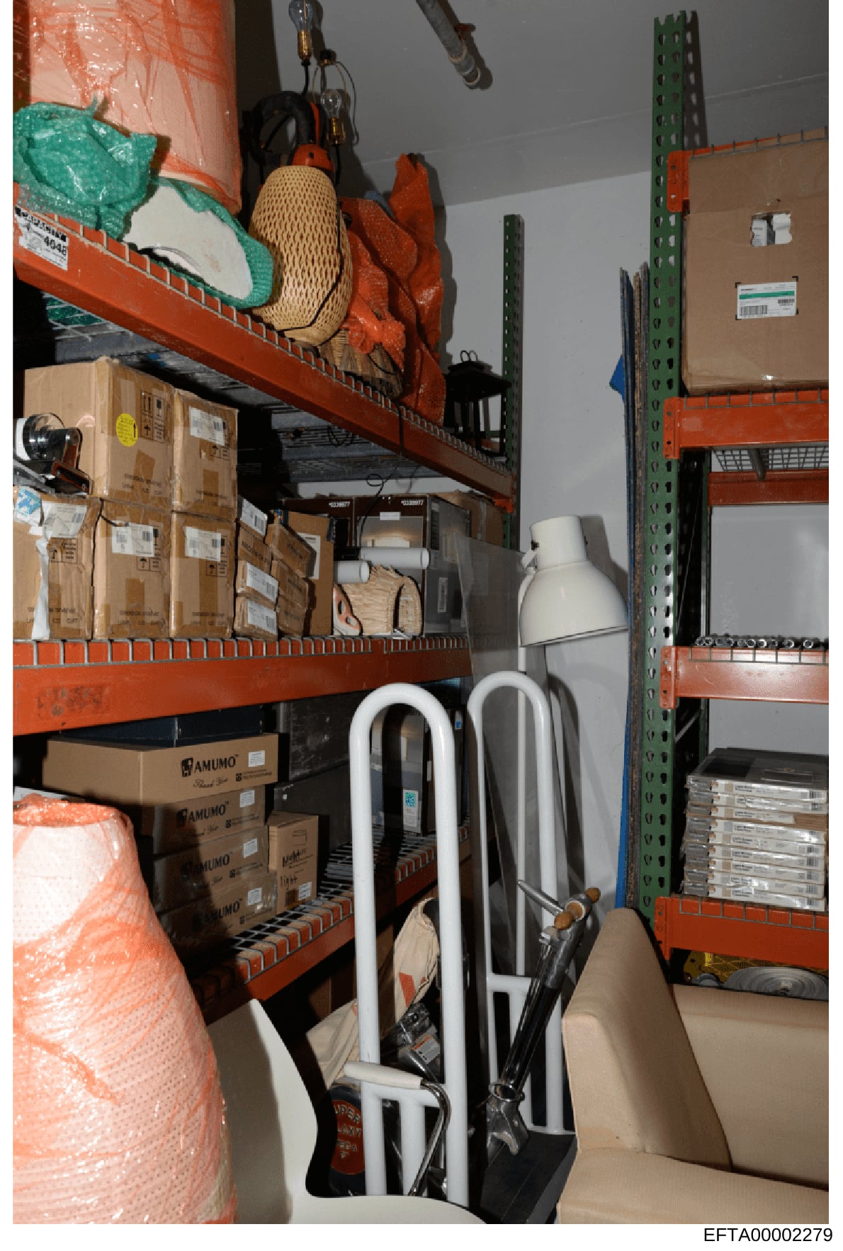 This is a photograph of a storage or warehouse space containing multiple cardboard boxes on industrial metal shelving, various equipment including rolled materials, and miscellaneous items. The image appears to be evidence documentation from the Epst
