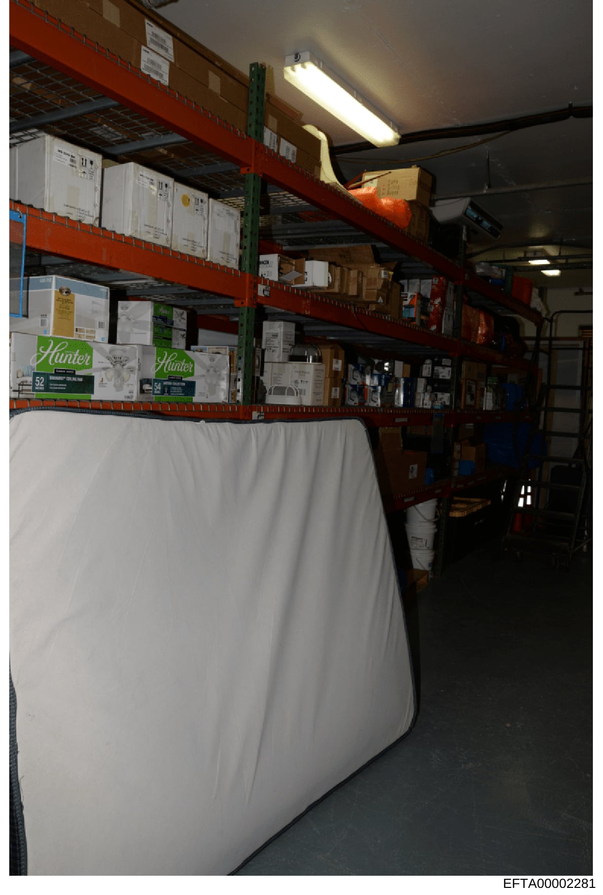 This is a photograph of an industrial storage warehouse showing red metal shelving units loaded with various boxed items, including Hunter brand products visible on the lower shelf. The image appears to be evidence documentation from a property searc