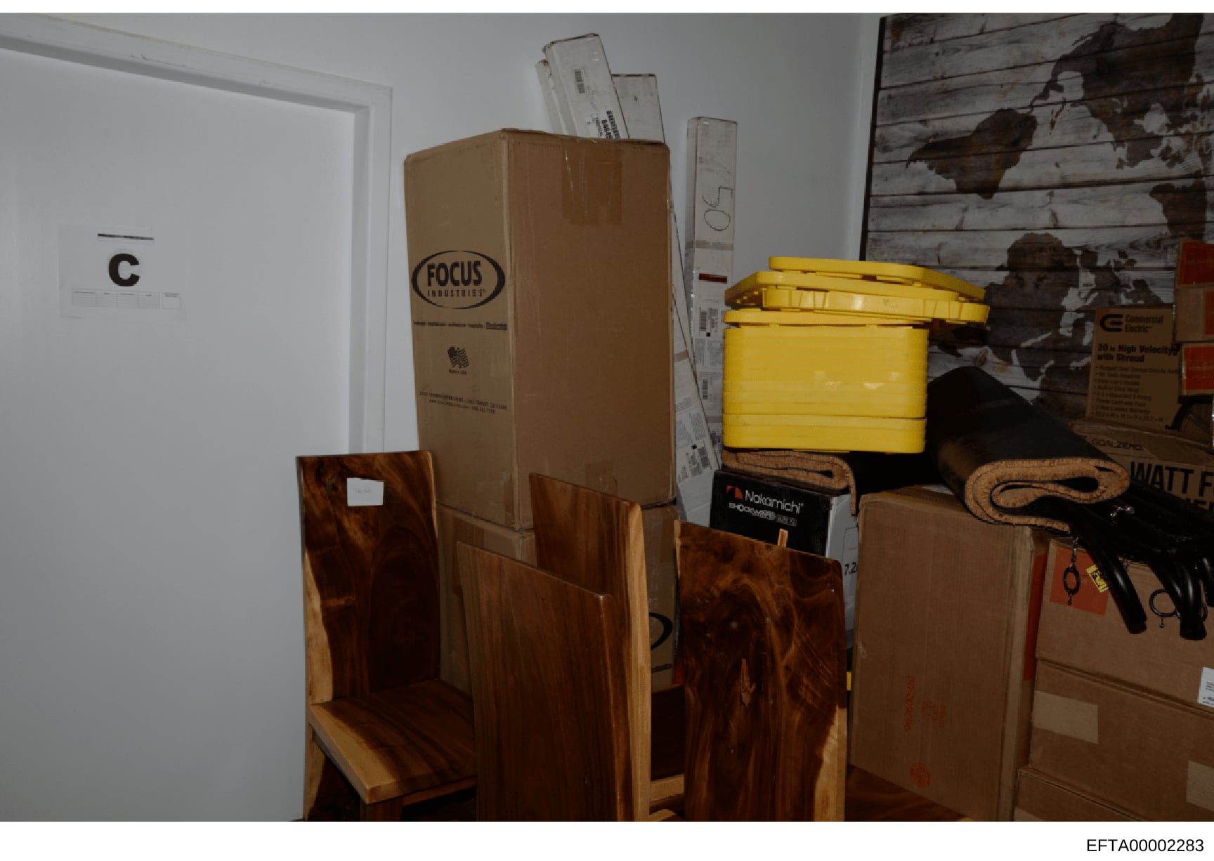 This is a photograph of a storage or utility room showing various items including cardboard boxes, wooden furniture pieces, a yellow plastic container, tools, and other household materials. The image appears to be documentation of property contents,