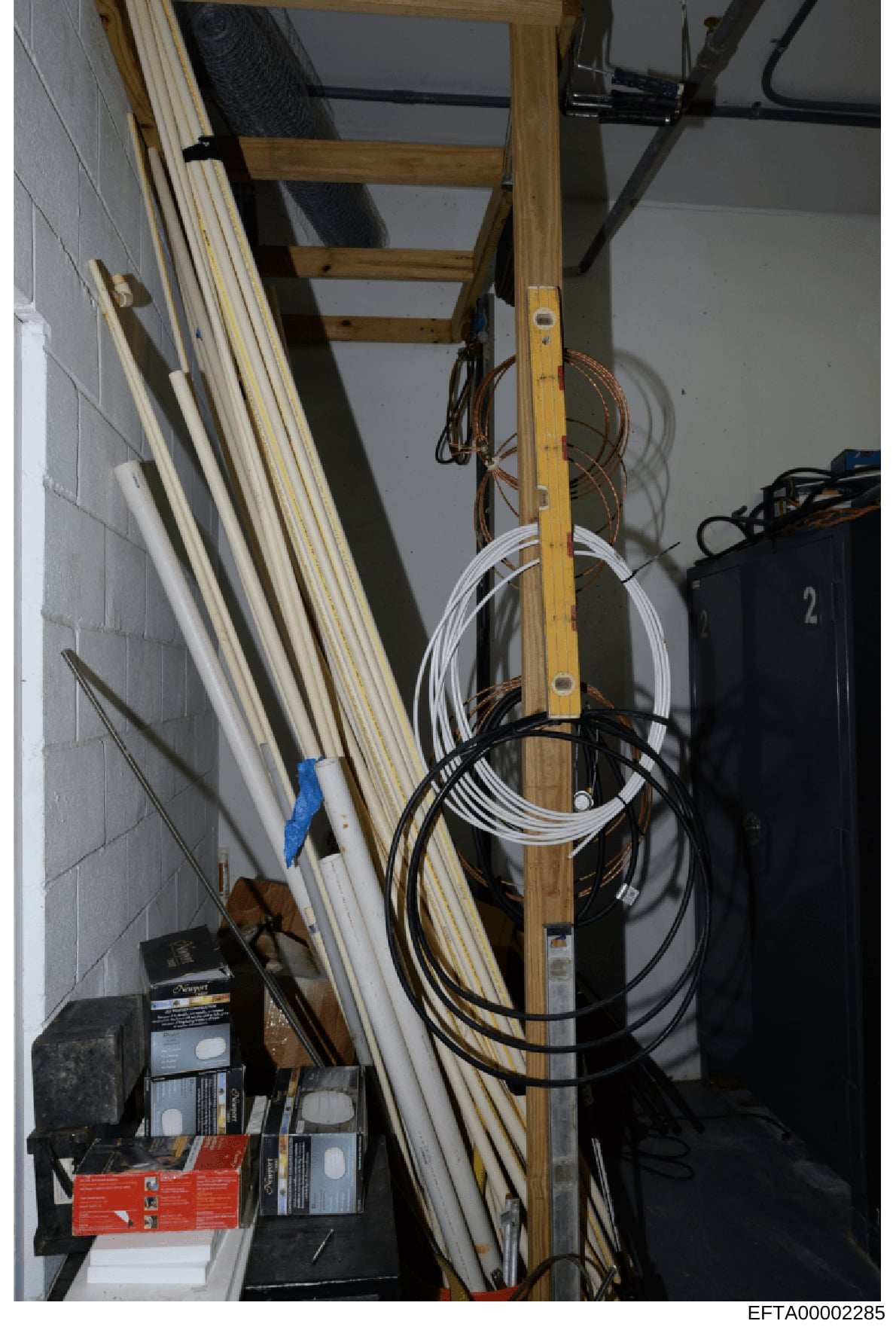 This photograph depicts an interior storage or utility space, likely a basement or closet area, containing building materials, electrical cables, extension cords, and hardware supplies. The image shows wooden ladder segments leaning against a wall, v