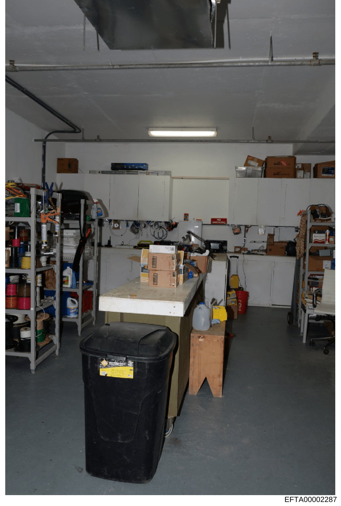 This is a photograph of an industrial or residential garage/storage space containing various tools, equipment, and supplies organized on shelves and work surfaces. The image shows a work area with metal shelving units, cardboard boxes, a black waste