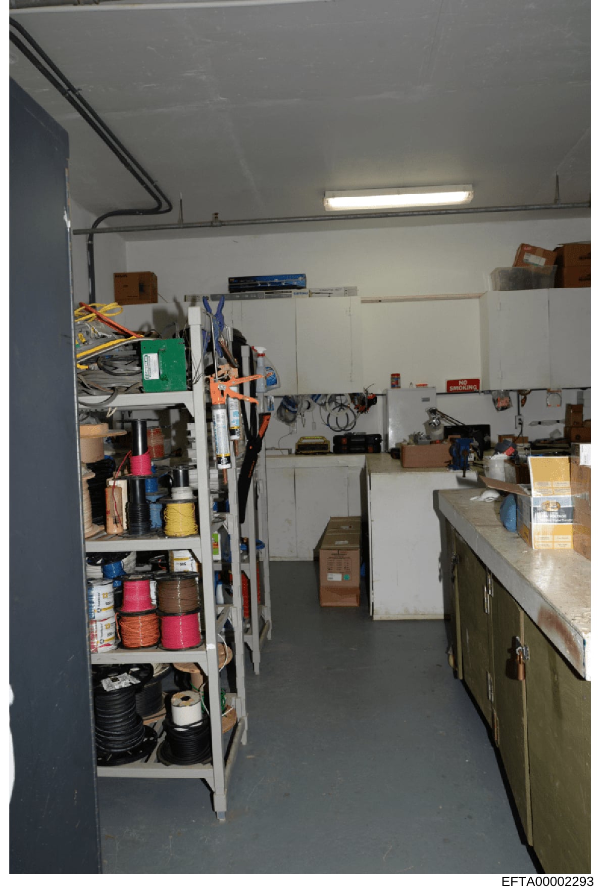 This photograph shows the interior of a utility or storage room, featuring white metal shelving units stocked with various electrical components, tools, supplies, and equipment. The room contains organized storage of cables, spools of wire or rope in