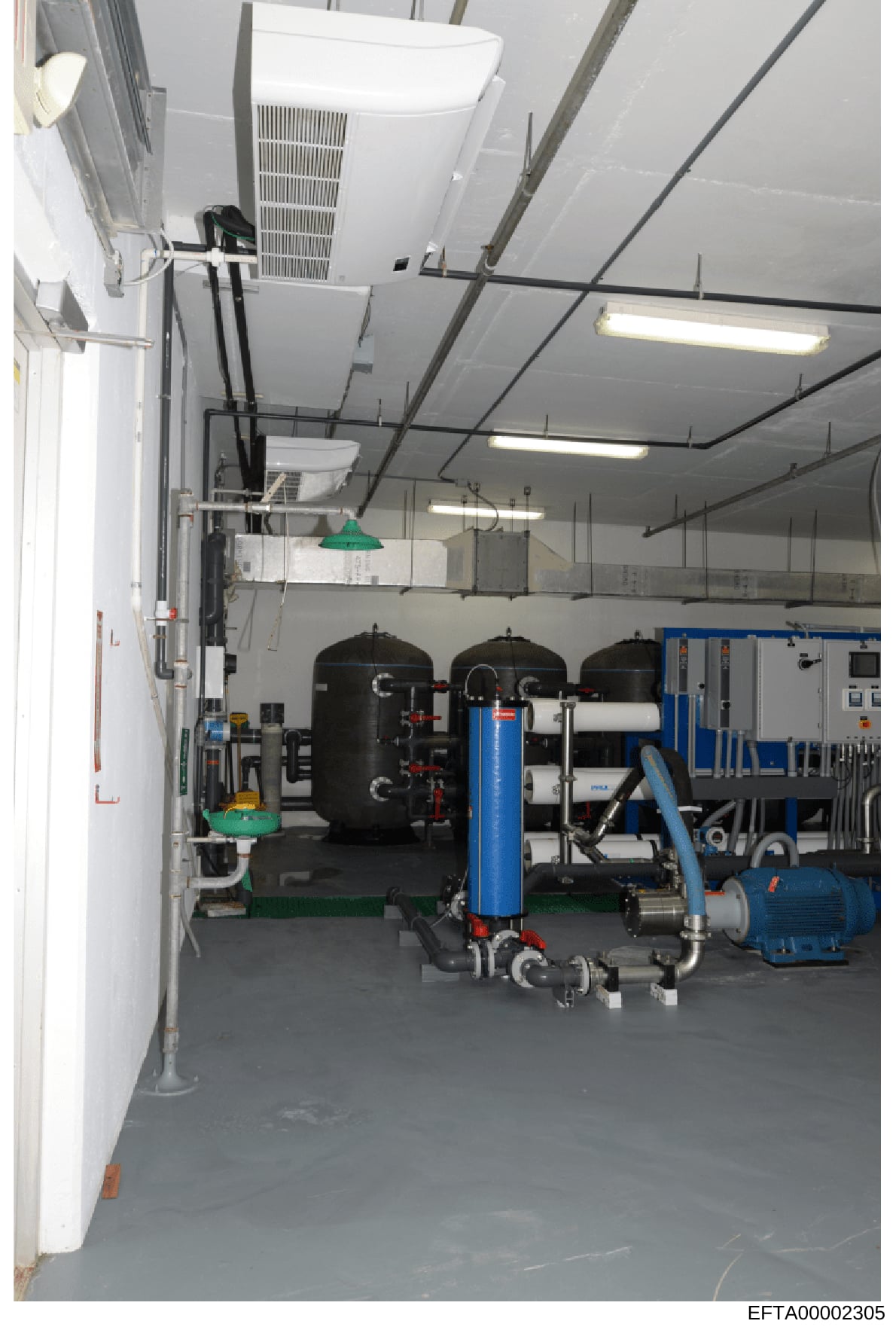 This photograph depicts an interior mechanical or equipment room containing industrial-scale water filtration and treatment systems. The space shows multiple black pressure tanks, a large blue cylindrical filter, pumps, piping infrastructure, and HVA