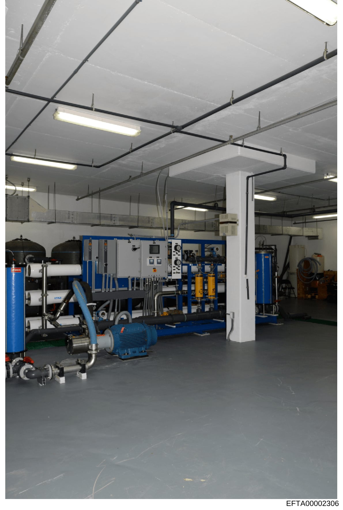 This photograph shows an industrial or commercial facility interior with specialized water treatment or processing equipment. The space contains blue-colored machinery, control panels, filtration systems, and various technical equipment arranged in a