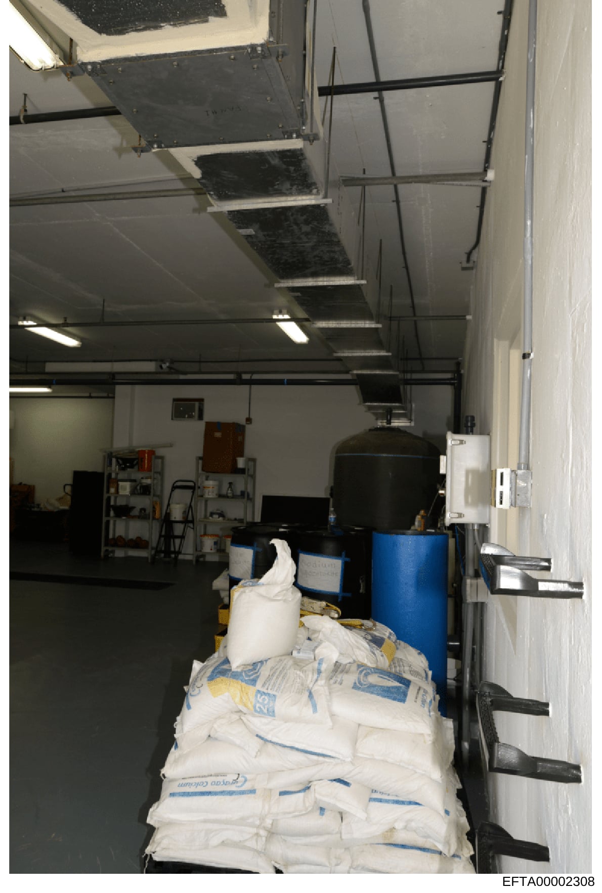 This photograph shows an interior storage or utility space, likely part of a larger facility, containing various supplies including large white bags (possibly containing bulk materials), blue drums/containers, shelving units, and HVAC equipment. The 