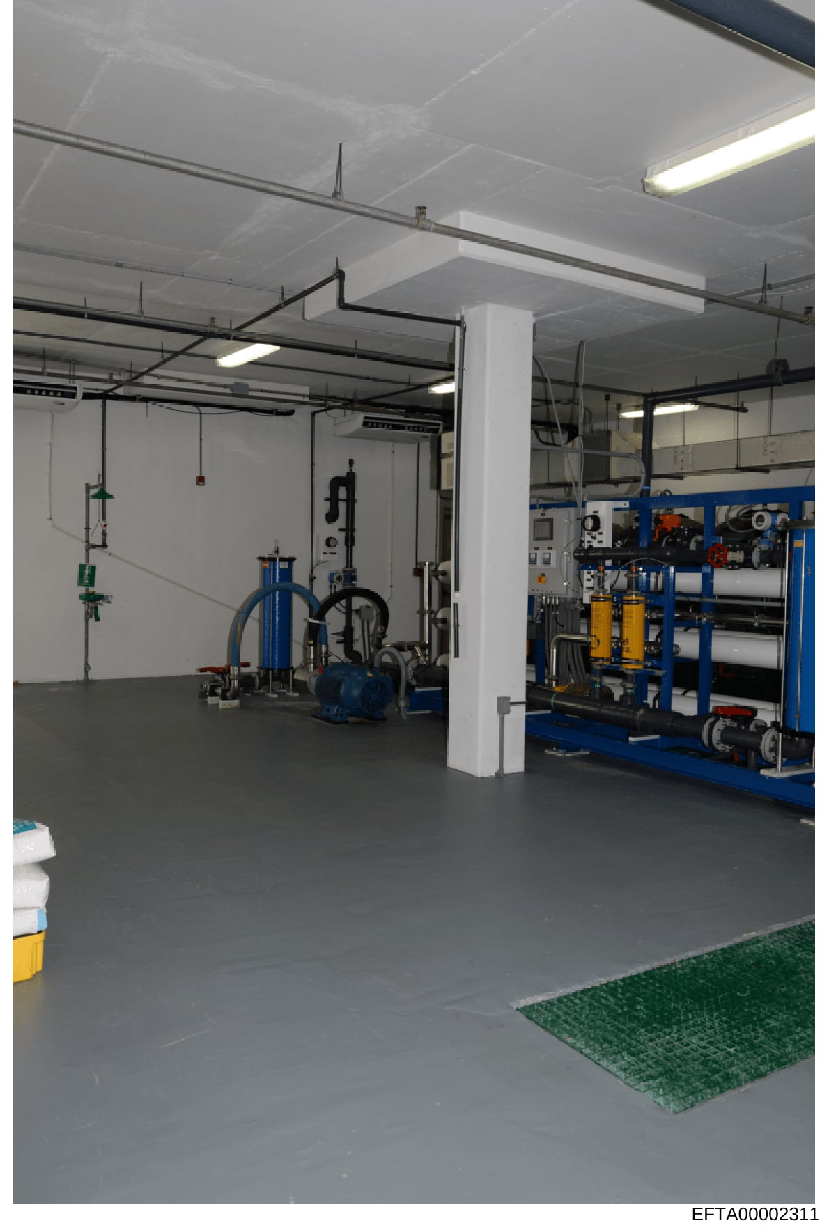 This photograph shows an interior view of an industrial or technical facility space. The room contains various mechanical equipment including blue hydraulic machinery, pumps, piping systems, and electrical control panels mounted on the walls. The spa