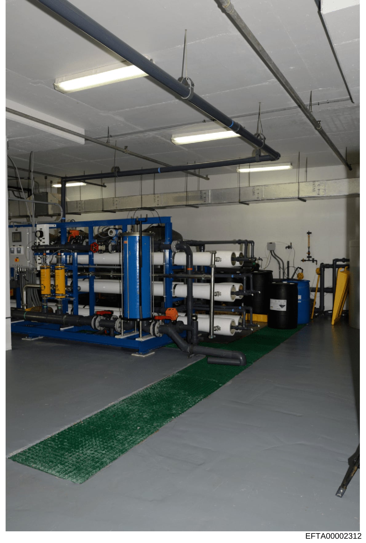 This photograph shows an industrial facility or manufacturing space containing large blue-framed equipment with cylindrical components, piping systems, and various tools. The space appears to be a technical or engineering facility with organized mach