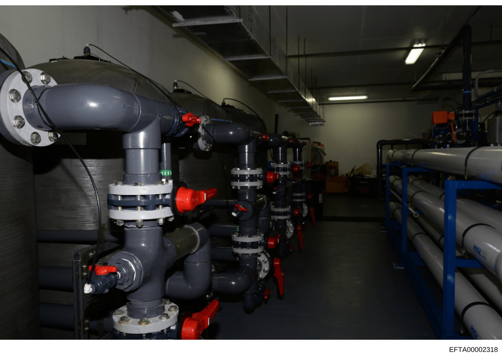 This is a photograph of an industrial facility showing multiple large dark gray/navy pipes with red valve controls and white flanged connections arranged in a warehouse or mechanical room setting. The equipment appears to be part of a water treatment