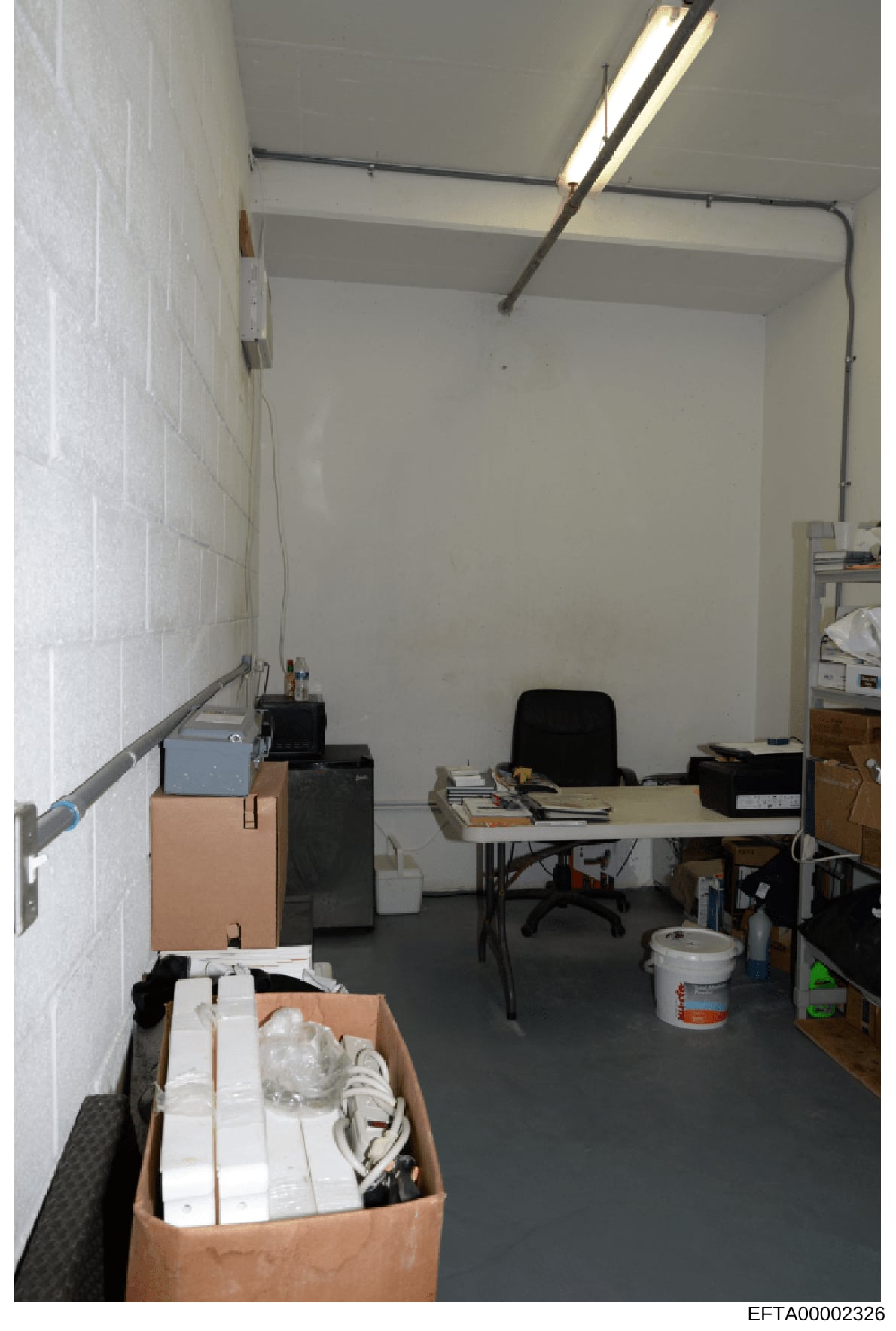 This is an evidence photograph showing an interior basement room or office space containing cardboard boxes with various materials, office furniture including a desk and black office chair, fluorescent lighting, white painted cinder block walls, and 